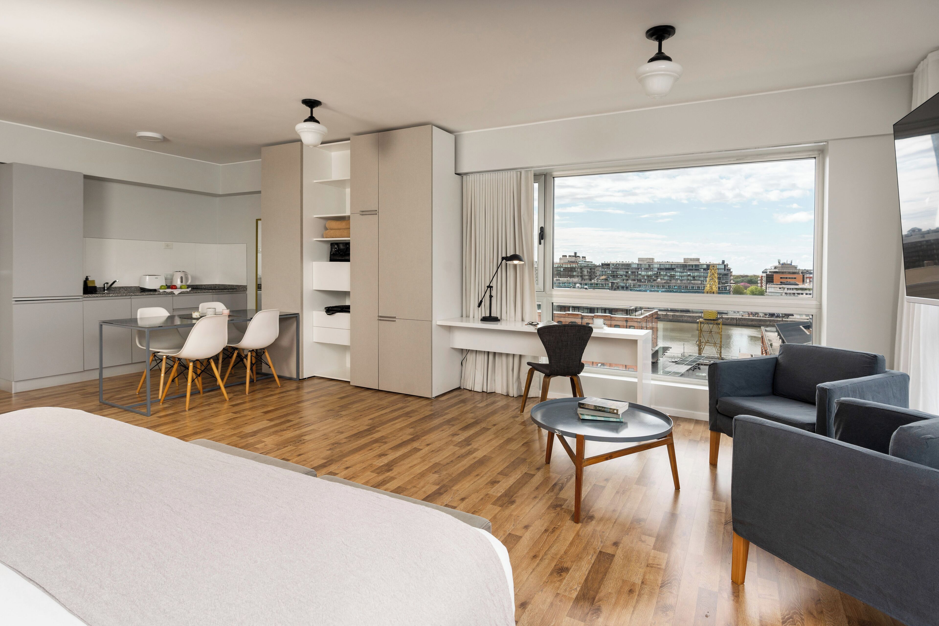 A modern hotel room with a large bed, a dining area with chairs, a desk with a chair, and a view of the city through large windows.