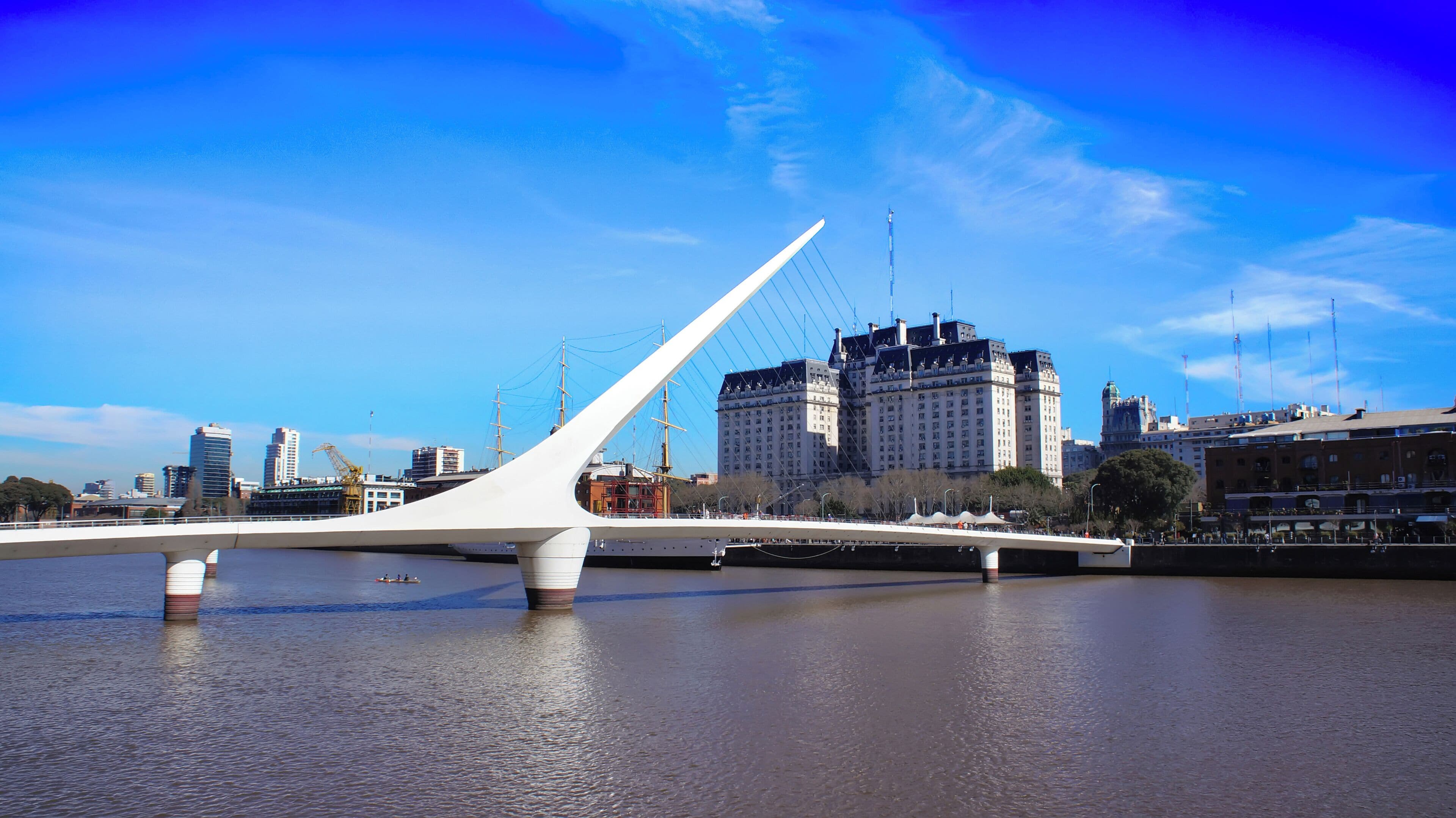 Uma ponte moderna com design branco semelhante a um veleiro, atravessando um rio, com um horizonte urbano ao fundo.