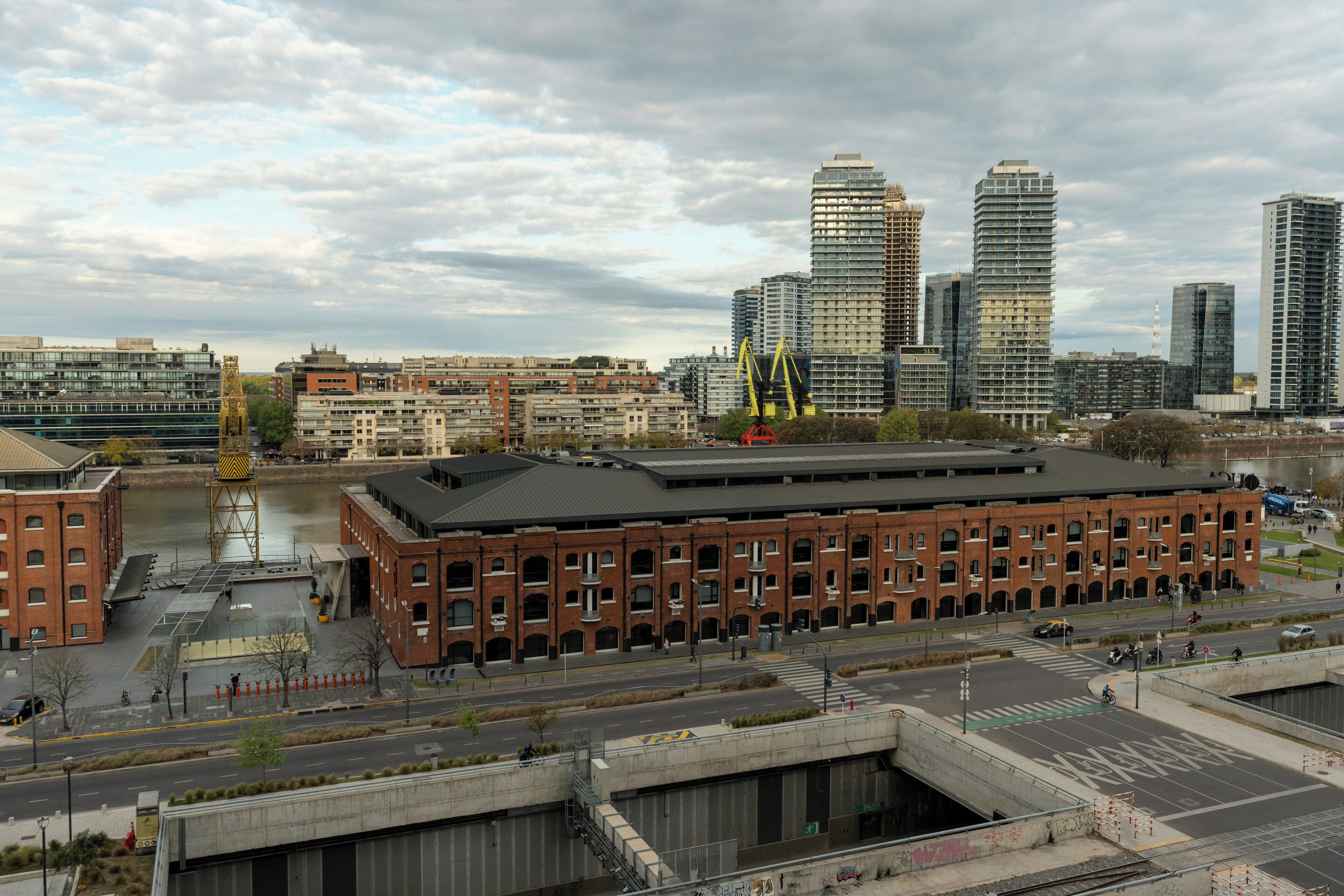 A large brick building with multiple floors, surrounded by modern high-rise buildings and a river.