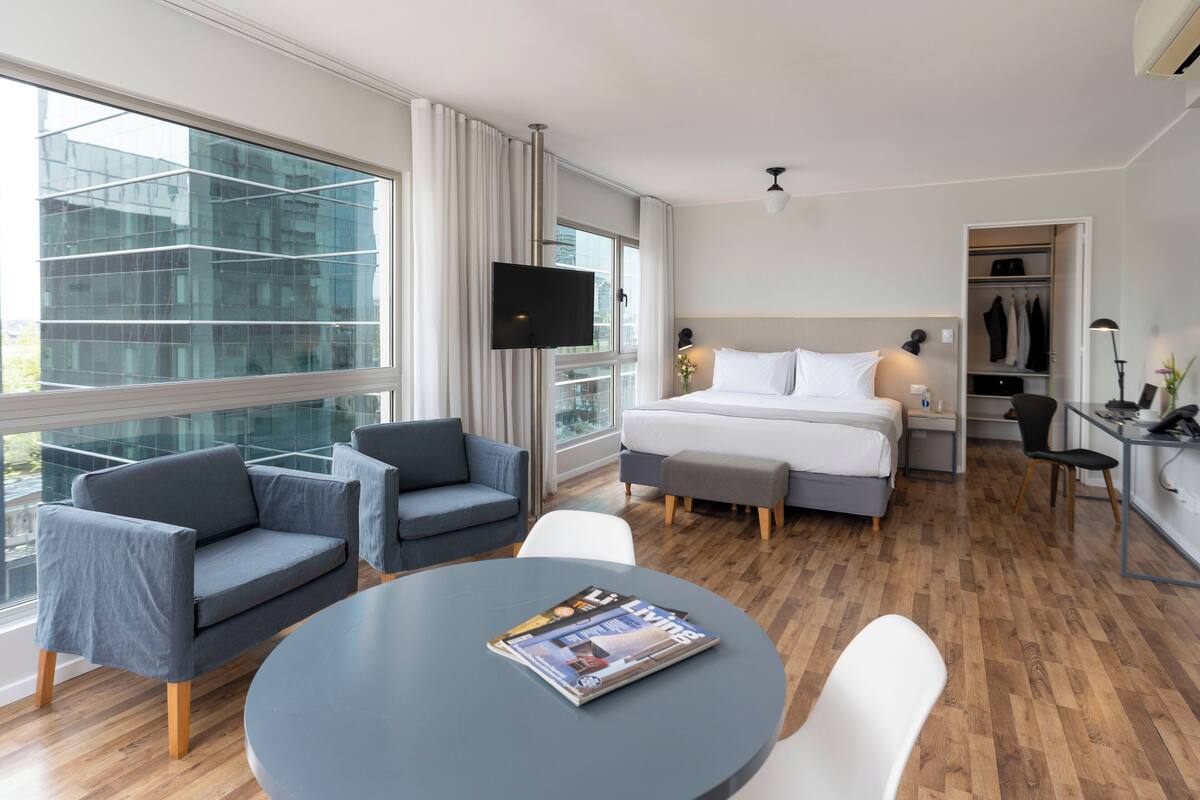 A modern hotel room with a large window, a bed, a desk, and a seating area.