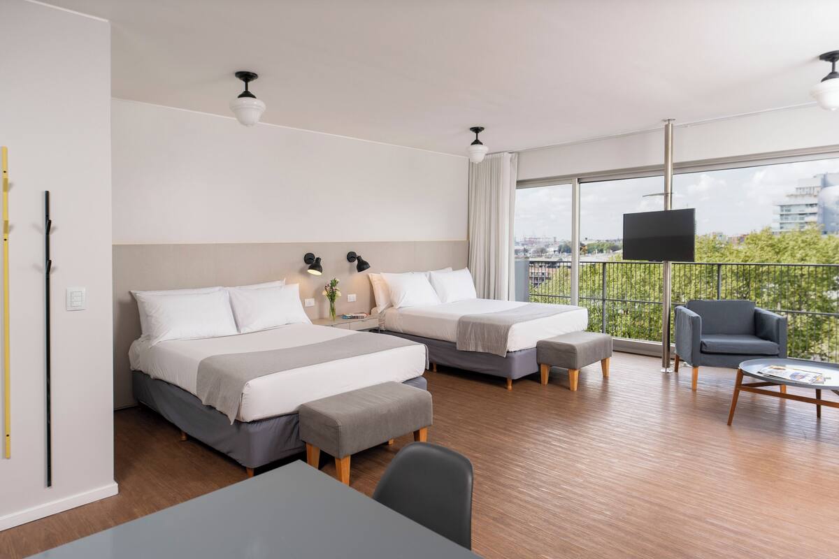 A modern hotel room with two beds, a balcony with a view, a TV, and a dining area.