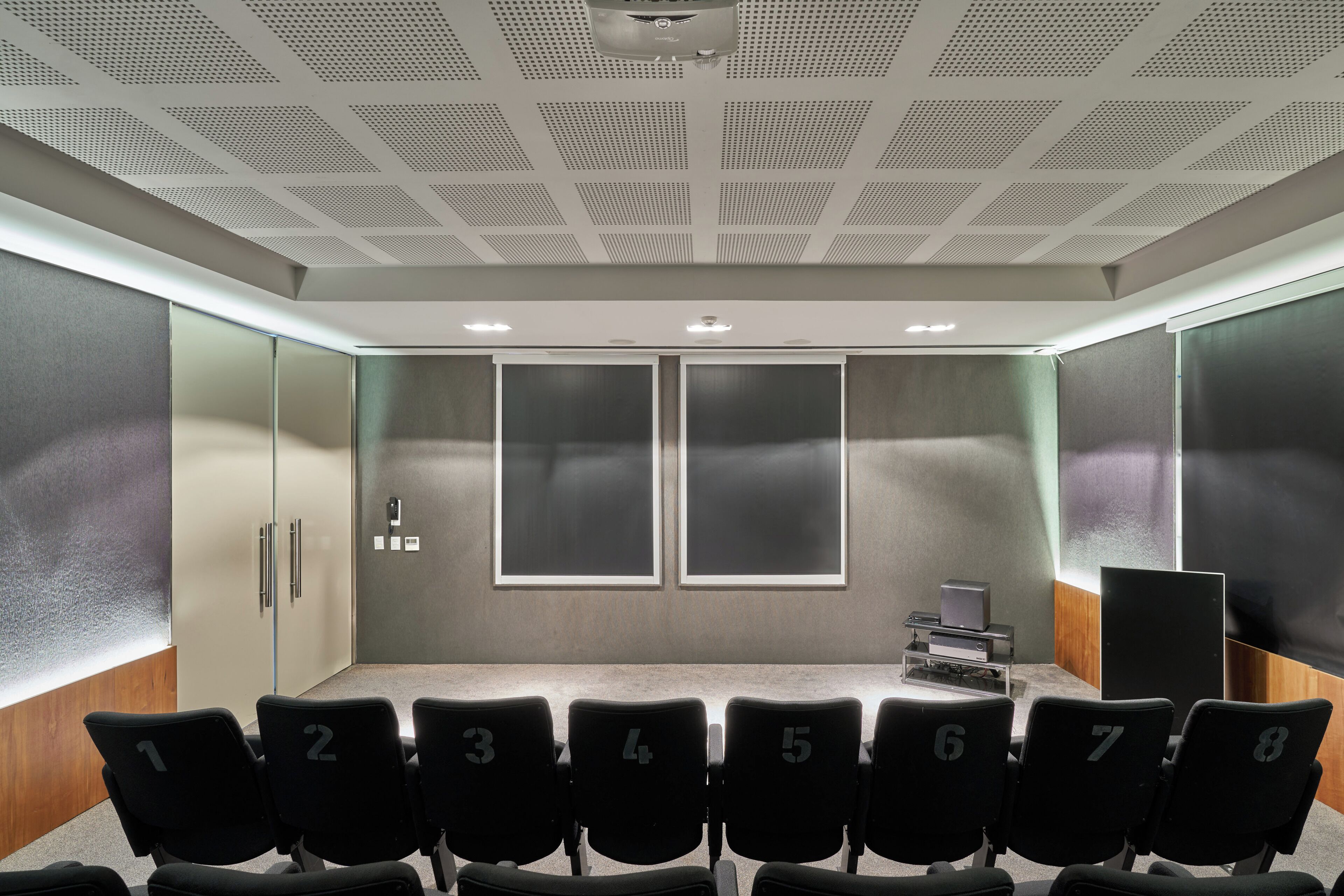 A conference room with a row of black chairs numbered 1-8, a podium, and a large screen on the right.