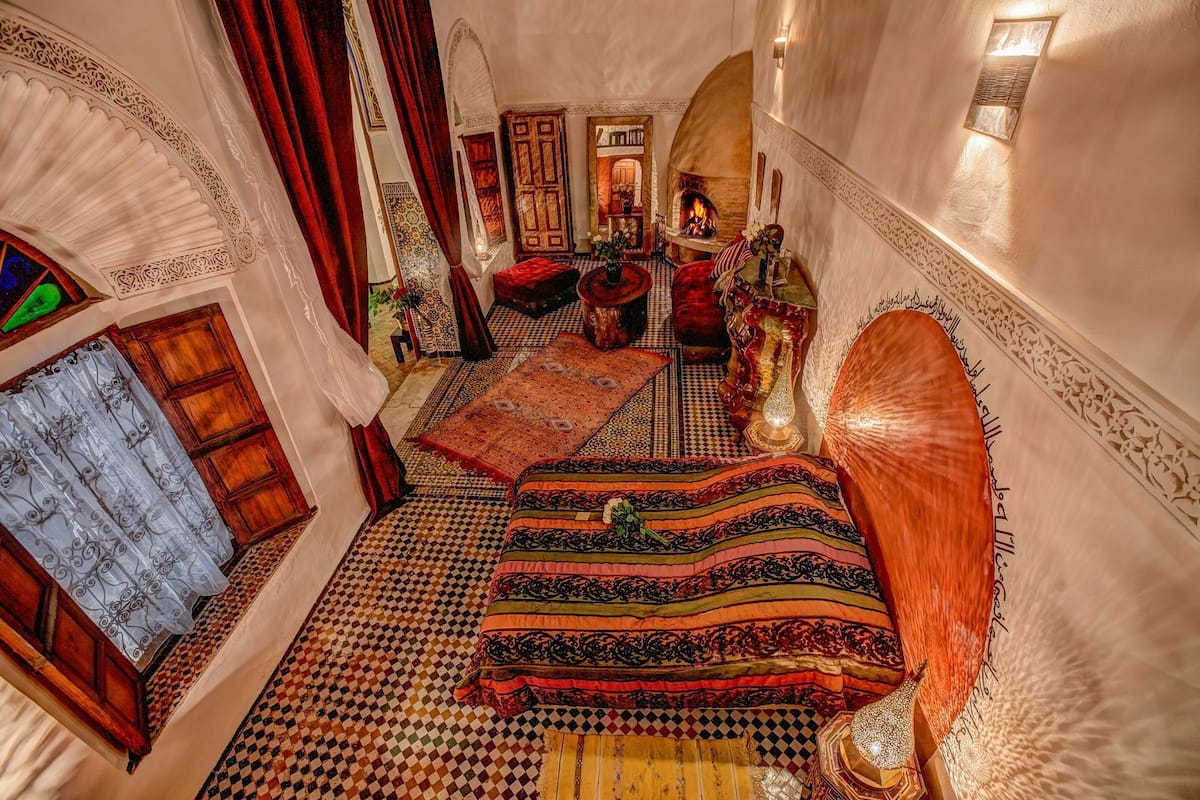 Quarto com piso xadrez, cama com colcha estampada, lareira e detalhes decorativos nas paredes.