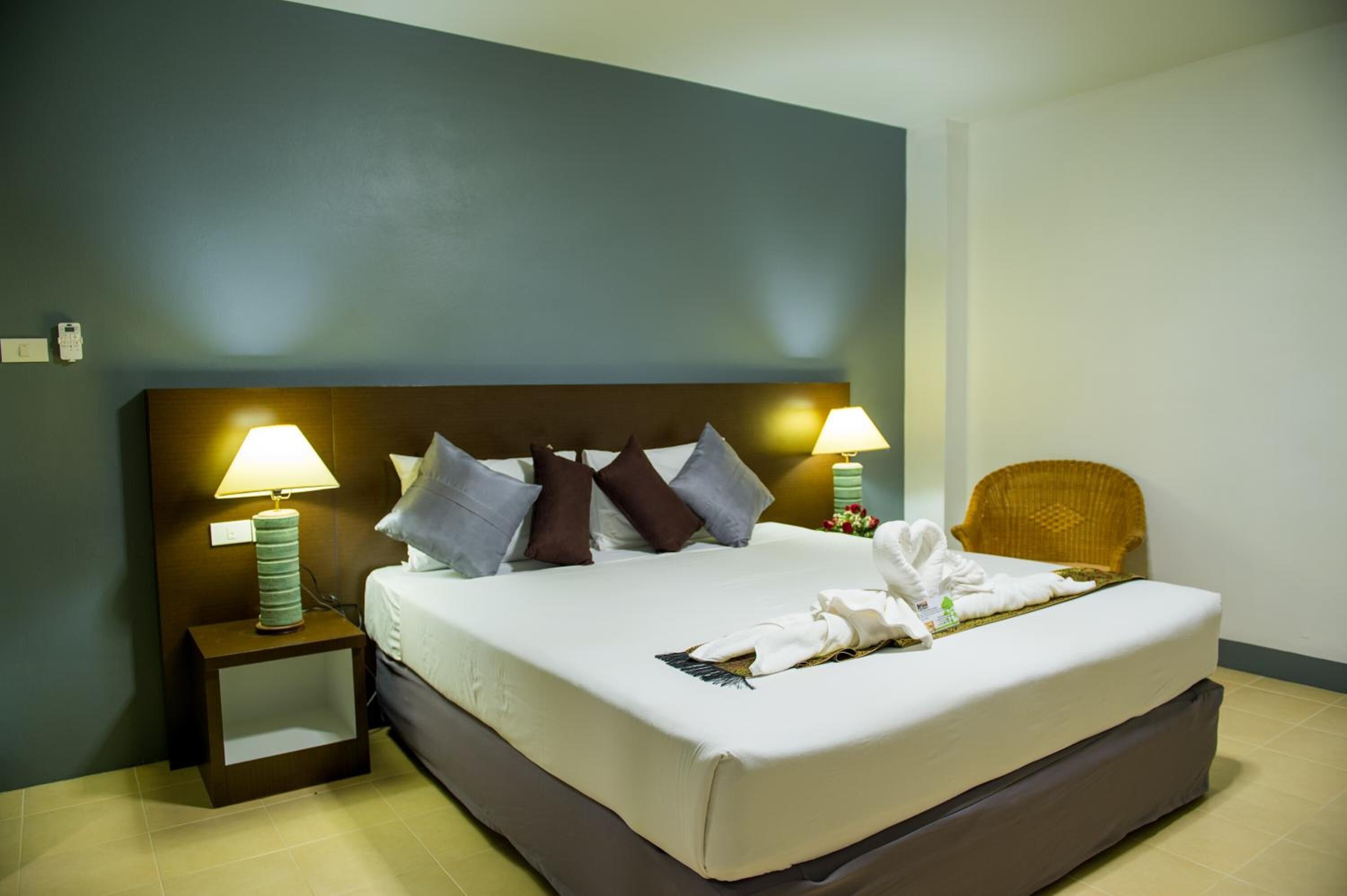 A neatly made bed with a headboard, two bedside lamps, a wicker chair, and a small nightstand with a lamp.