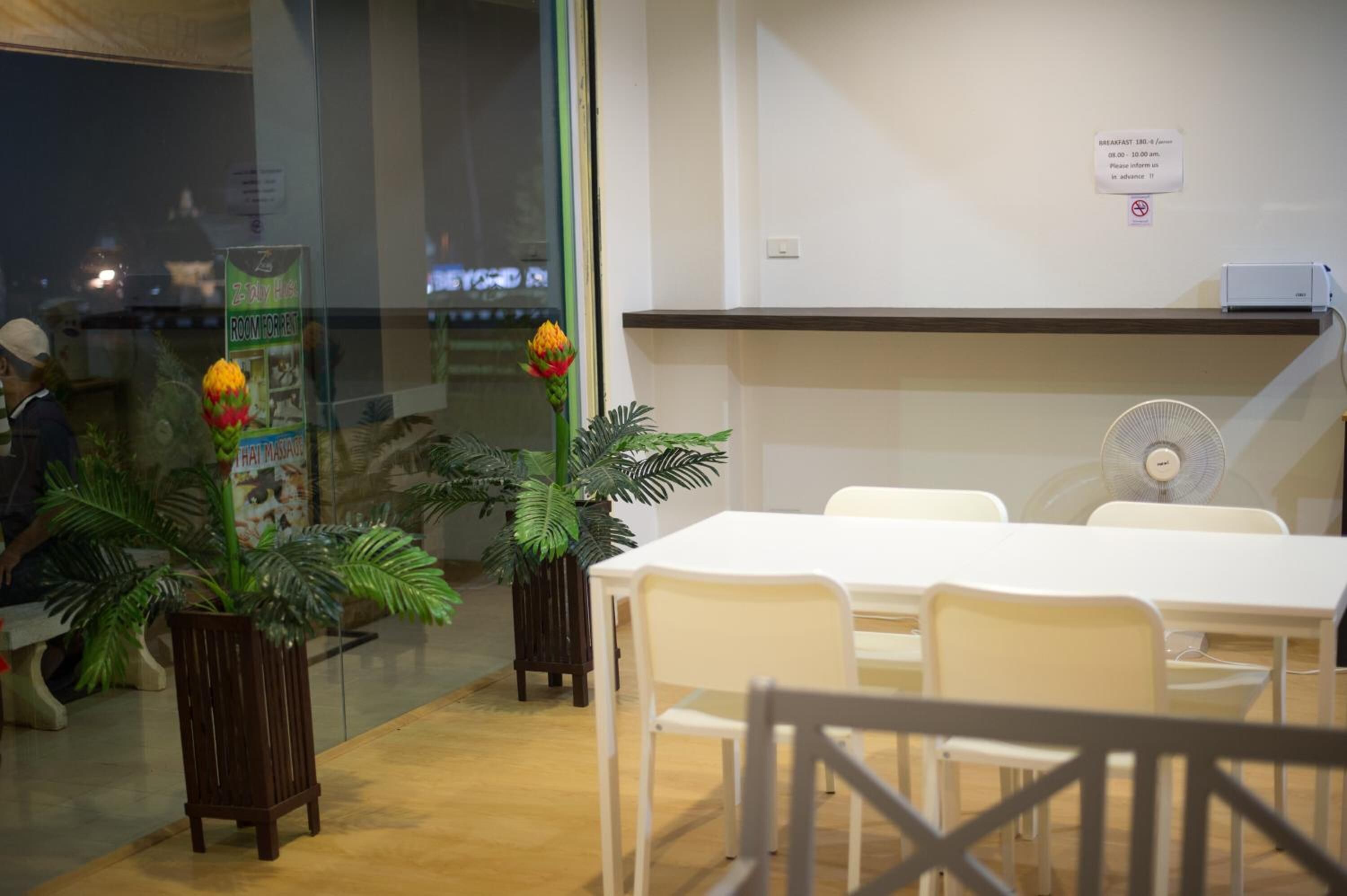 Un salón de conferencias con mesas y sillas blancas, un ventilador de pie y plantas en macetas.