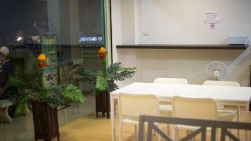 Un salón de conferencias con mesas y sillas blancas, un ventilador de pie y plantas en macetas.