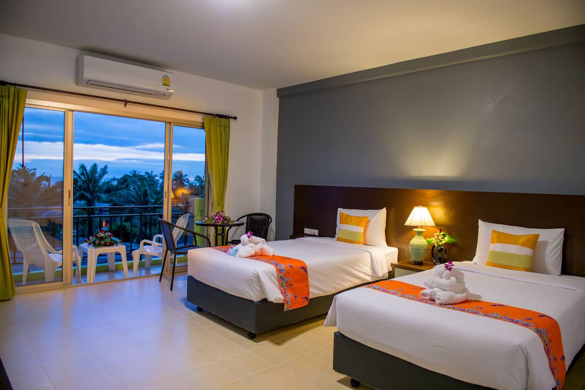 A hotel room with two beds, a balcony with chairs, a table with flowers, and a nightstand with a lamp.