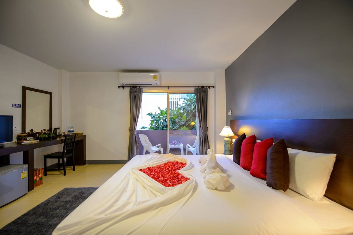 A hotel room with a bed, a desk, a chair, a TV, and a balcony with a view of greenery.