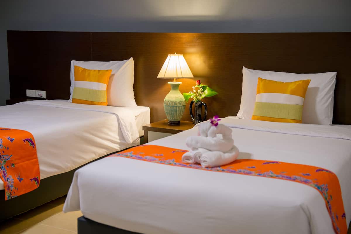 Two double beds with white linens and orange decorative borders, a bedside table with a lamp and a vase with flowers, and a towel on the bed.