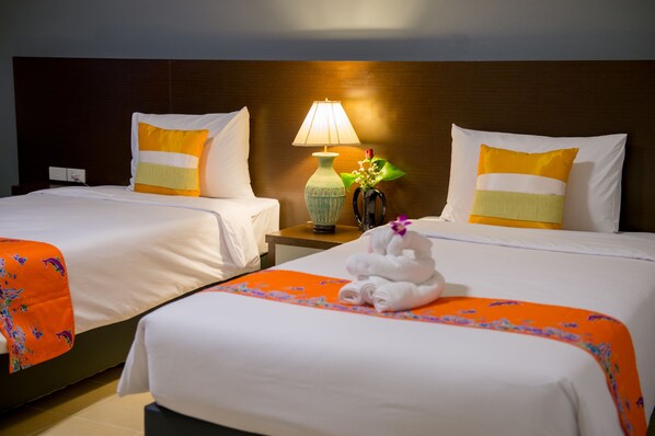 Duas camas de casal com roupa de cama branca e detalhes laranja decorativos, uma mesa de cabeceira com abajur e um vaso com flores, e uma toalha na cama.