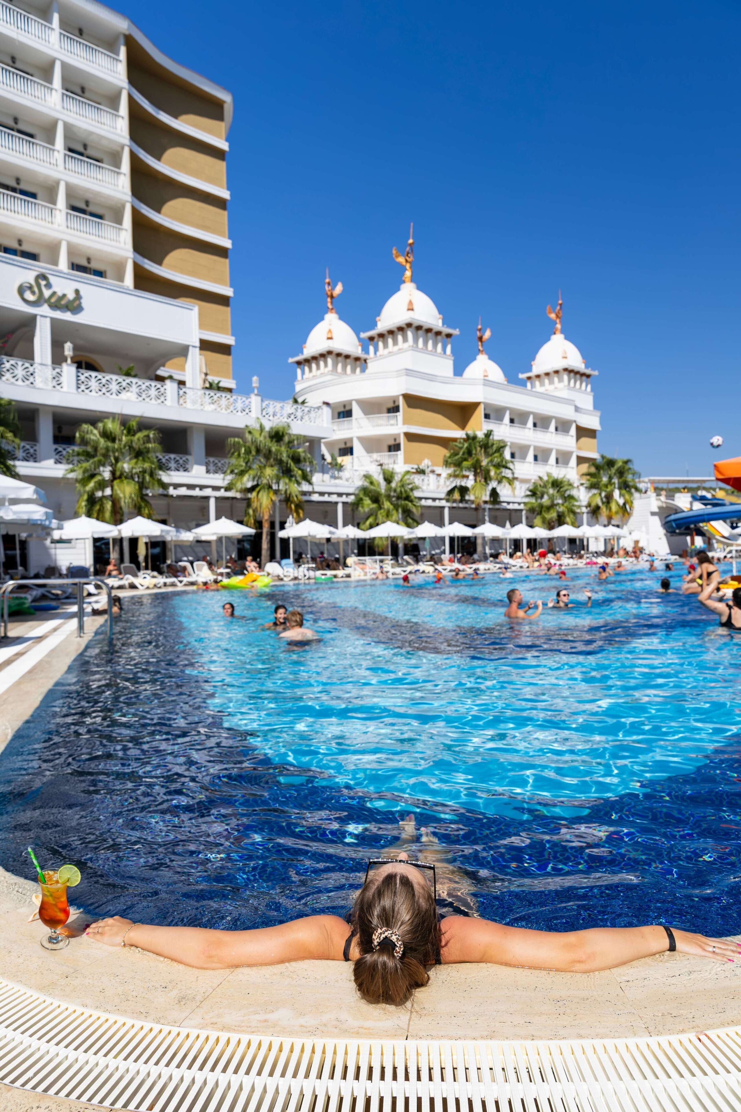 En swimmingpool med en person som kopplar av vid kanten, palmer och en hotellbyggnad med kupolformade konstruktioner i bakgrunden.