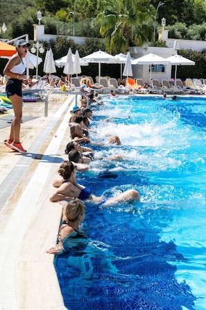 Ein Schwimmbecken mit Personen im Wasser und am Beckenrand, umgeben von Liegestühlen und Sonnenschirmen.