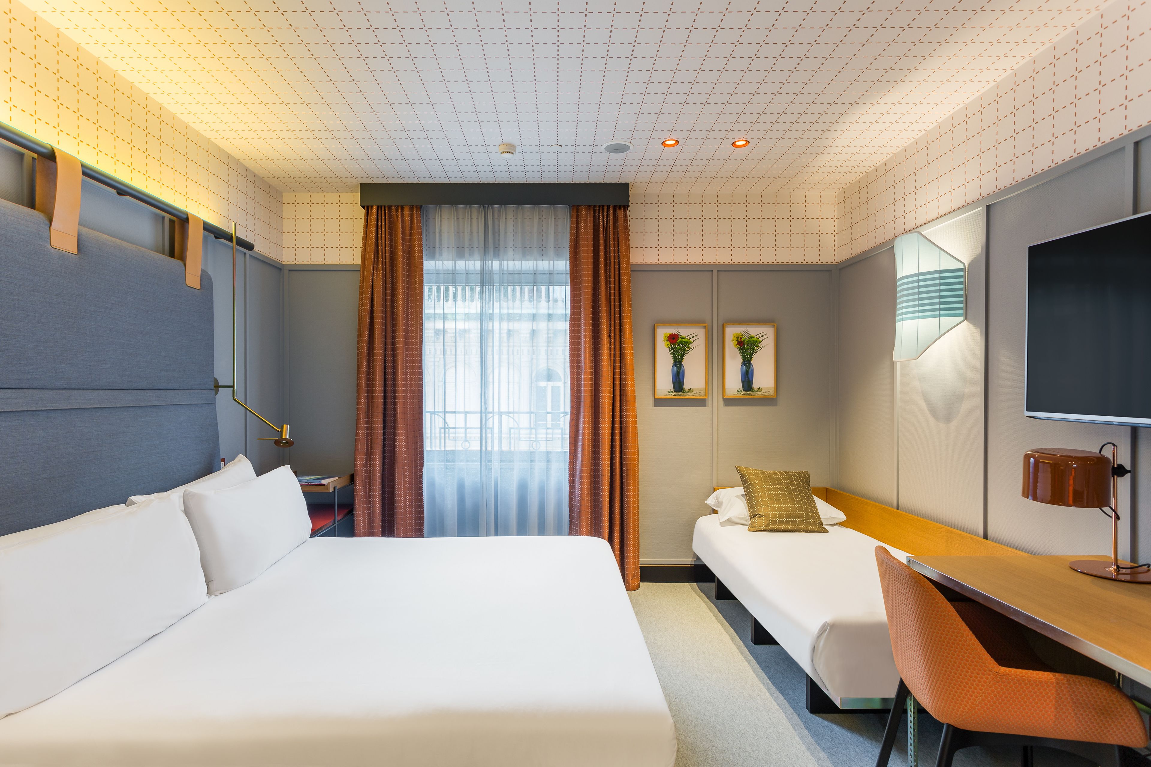 A hotel room with a large bed, a smaller bed, a desk with a chair, a TV, and two wall-mounted lamps.