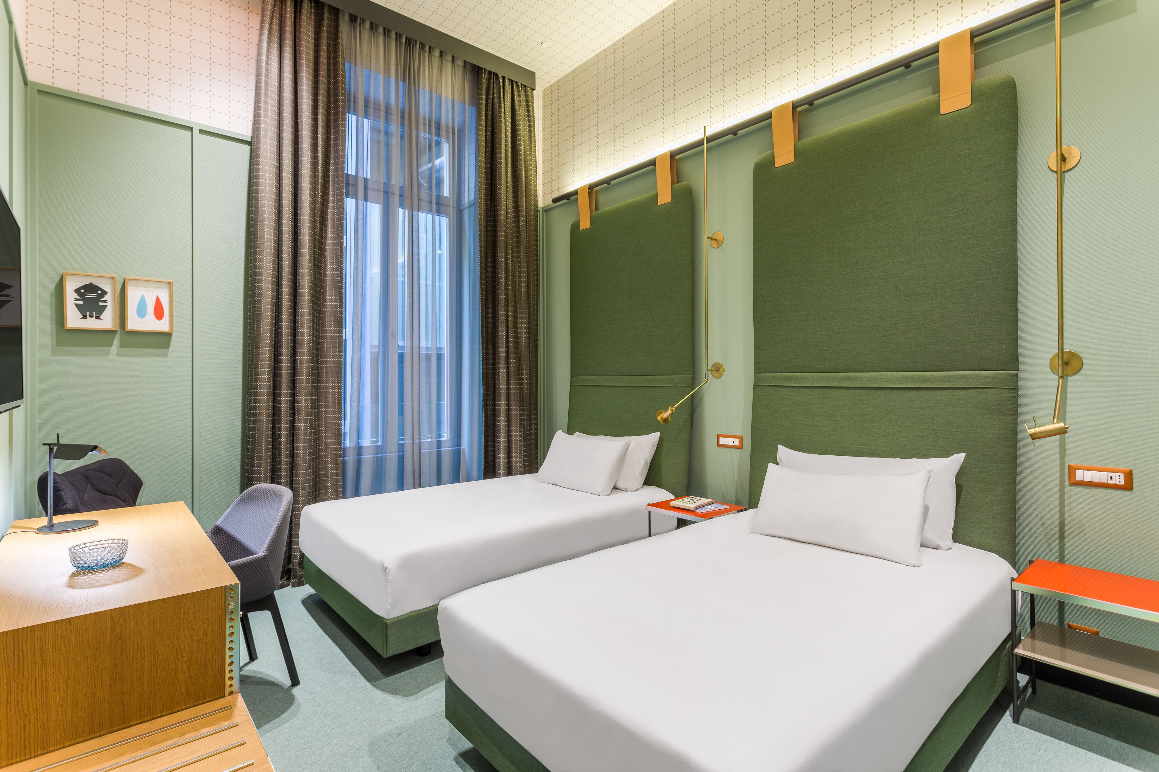 A hotel room with two single beds, a desk with a chair, a TV, and a window with curtains.