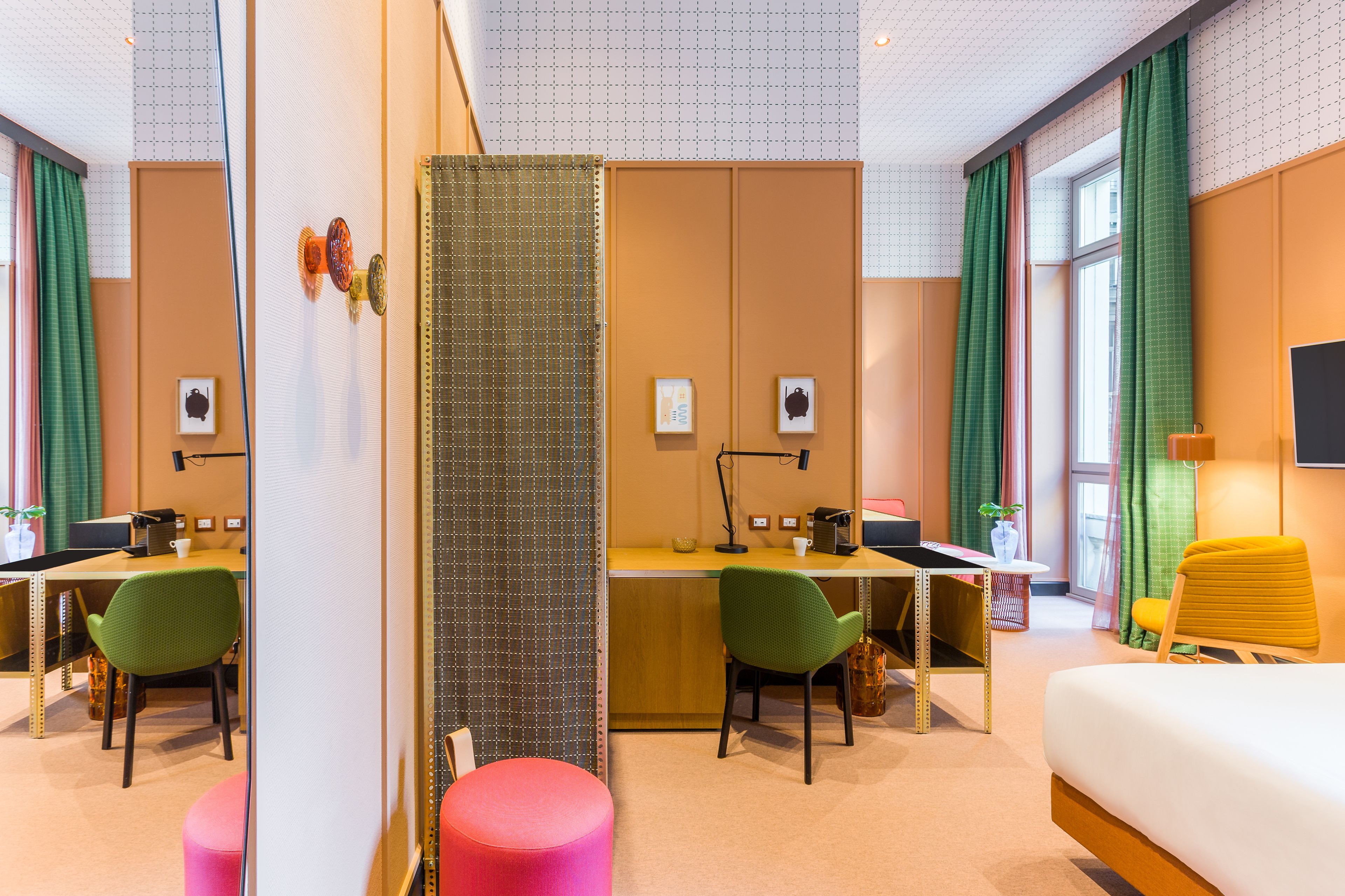 A hotel room with a bed, two desks with chairs, a small table with a lamp, a pink ottoman, and a large window with curtains.