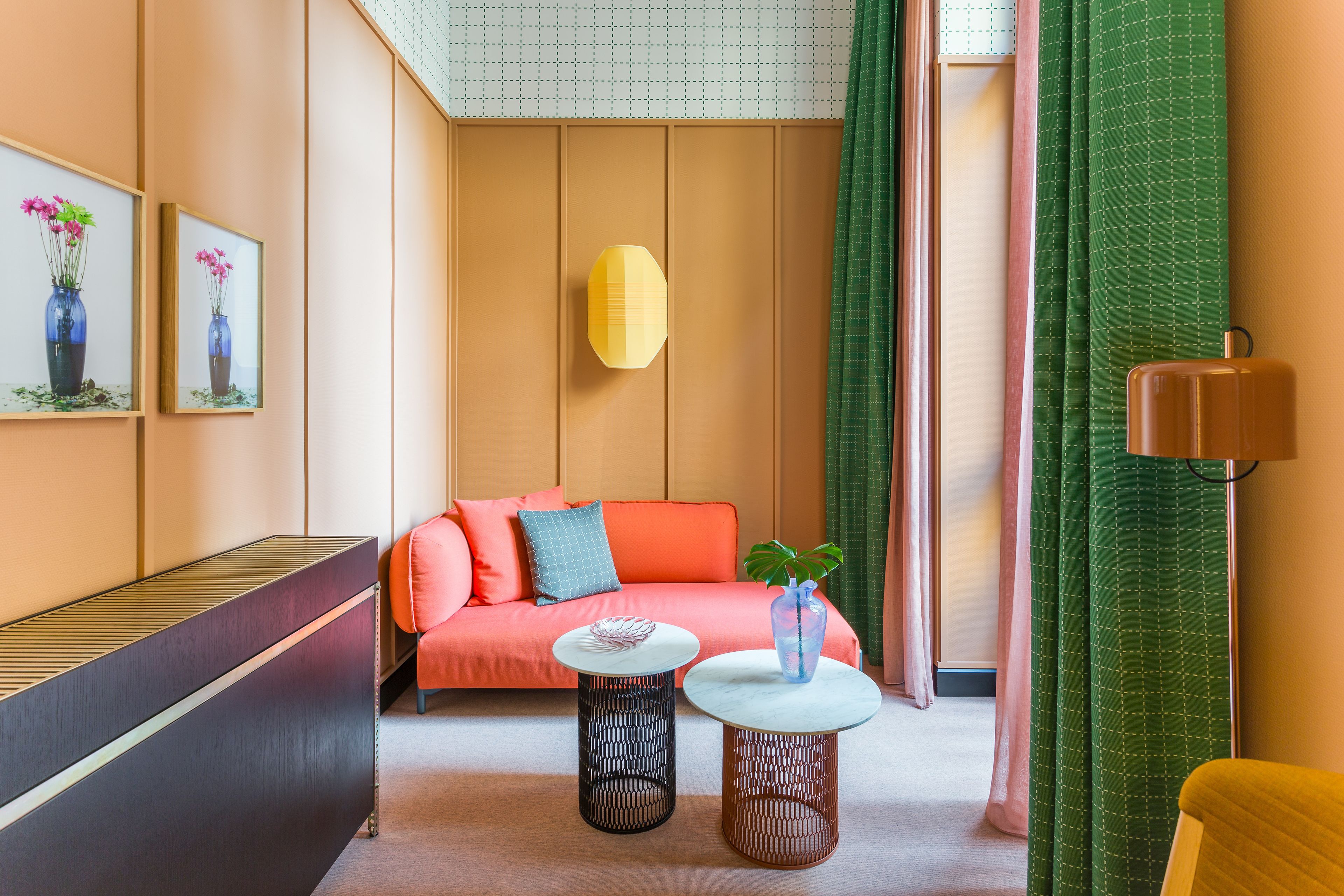 Ett modernt hotellrum med en soffa, två runda bord, en vas med blommor och inramade tavlor på väggen.