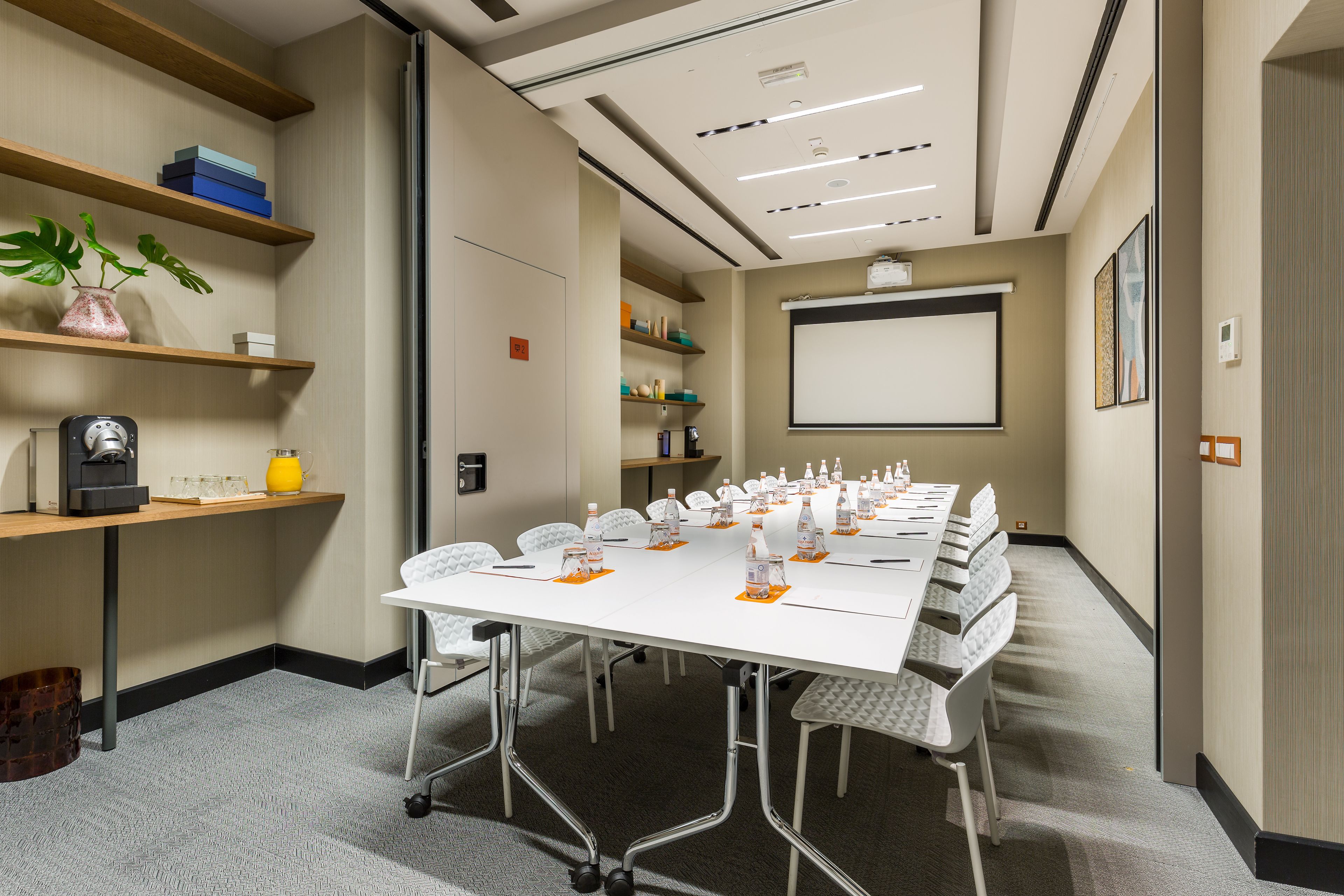 Uma sala de conferências com uma mesa comprida, cadeiras, uma tela de projetor e estantes com diversos objetos.