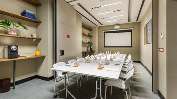 Uma sala de conferências com uma mesa comprida, cadeiras, uma tela de projetor e estantes com diversos objetos.