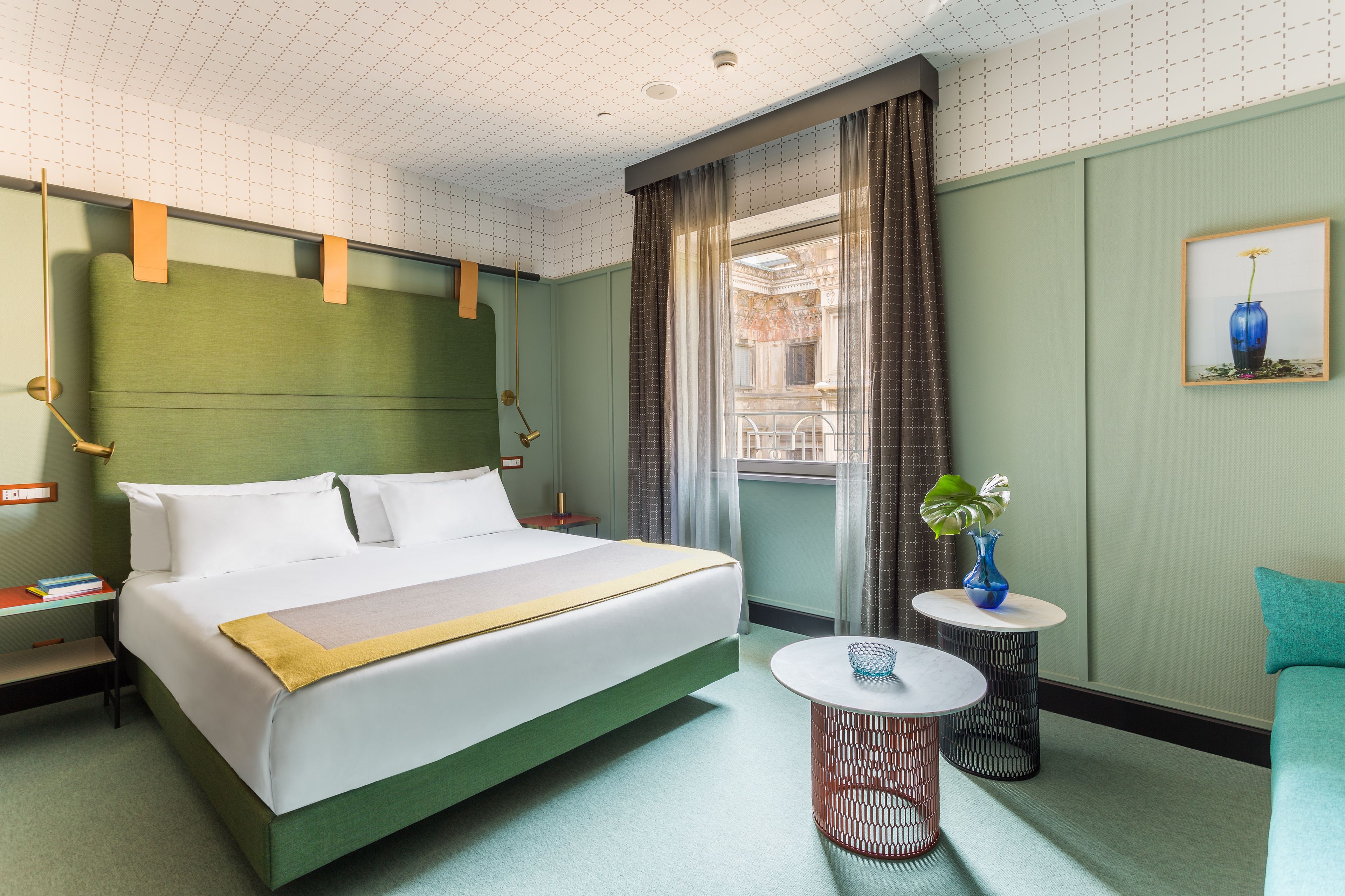 A hotel room with a large bed, a small round table, a chair, a window with curtains, and a framed picture on the wall.
