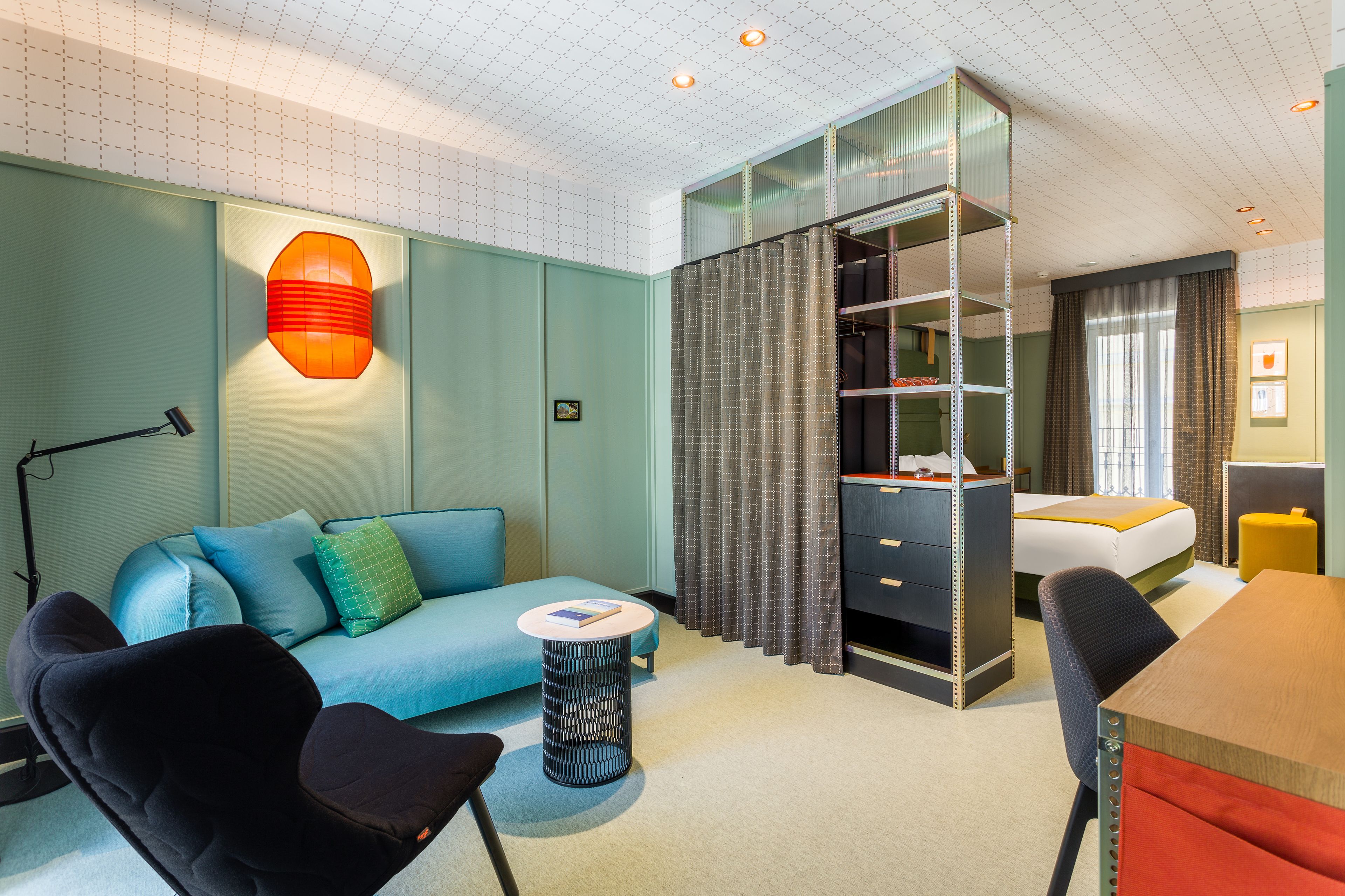 A hotel room with a blue sofa, a desk, a chair, a bookshelf, and a bed.