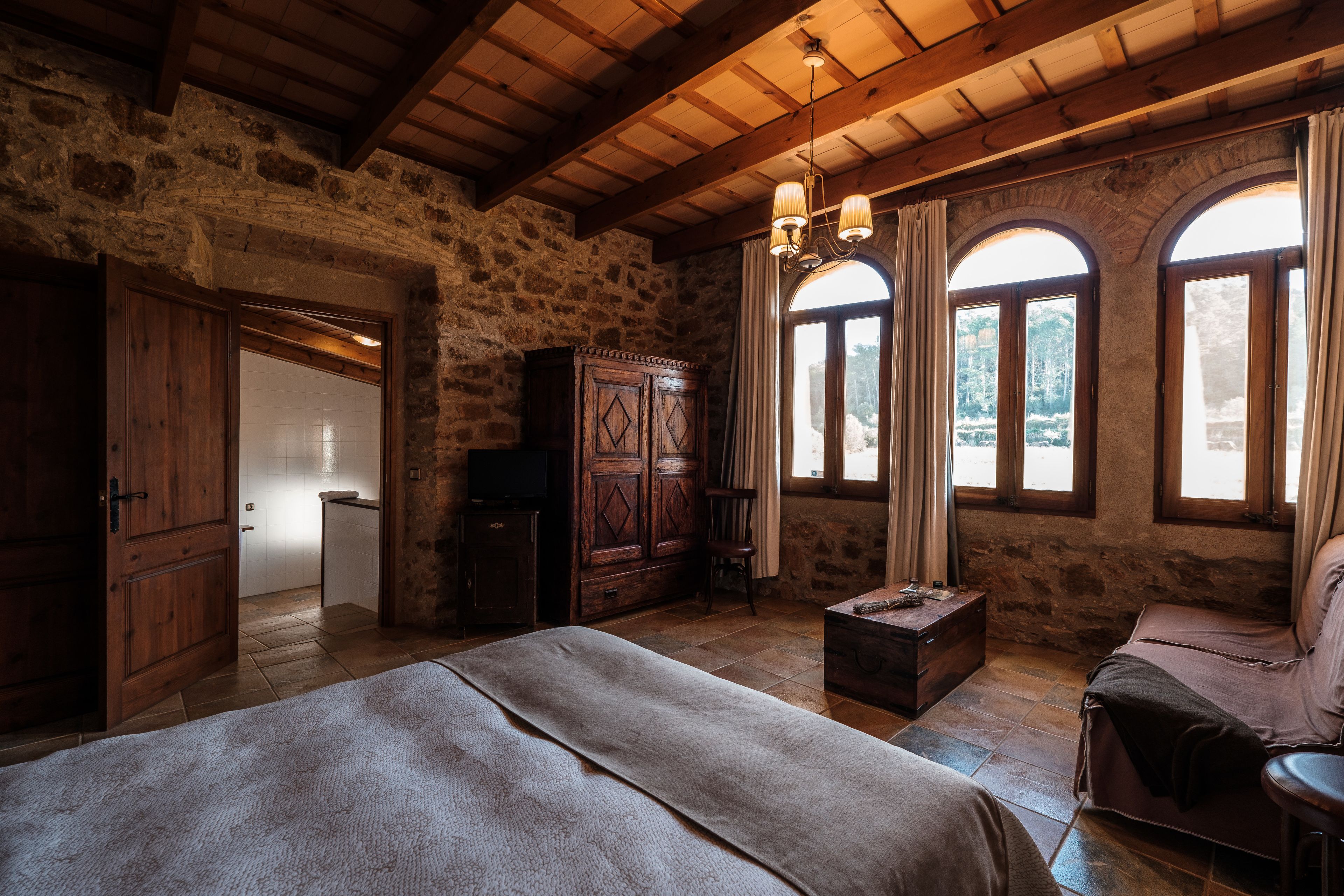 A rustic bedroom with stone walls, wooden beams, a large bed, a wooden wardrobe, and two windows with curtains.