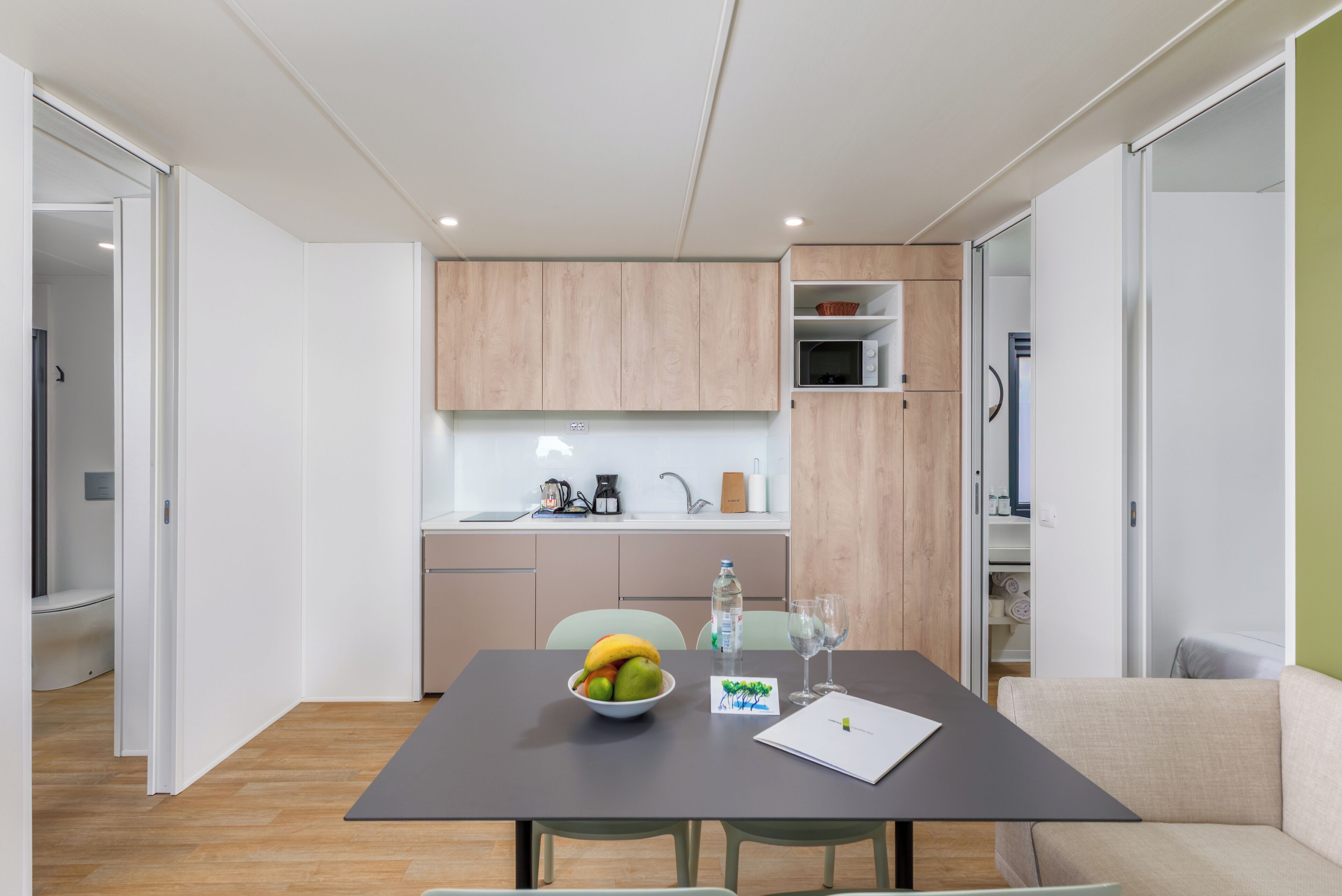 Une cuisine moderne avec des meubles en bois, un four à micro-ondes et un évier. On y trouve une table à manger avec un saladier rempli de fruits et un verre d’eau.