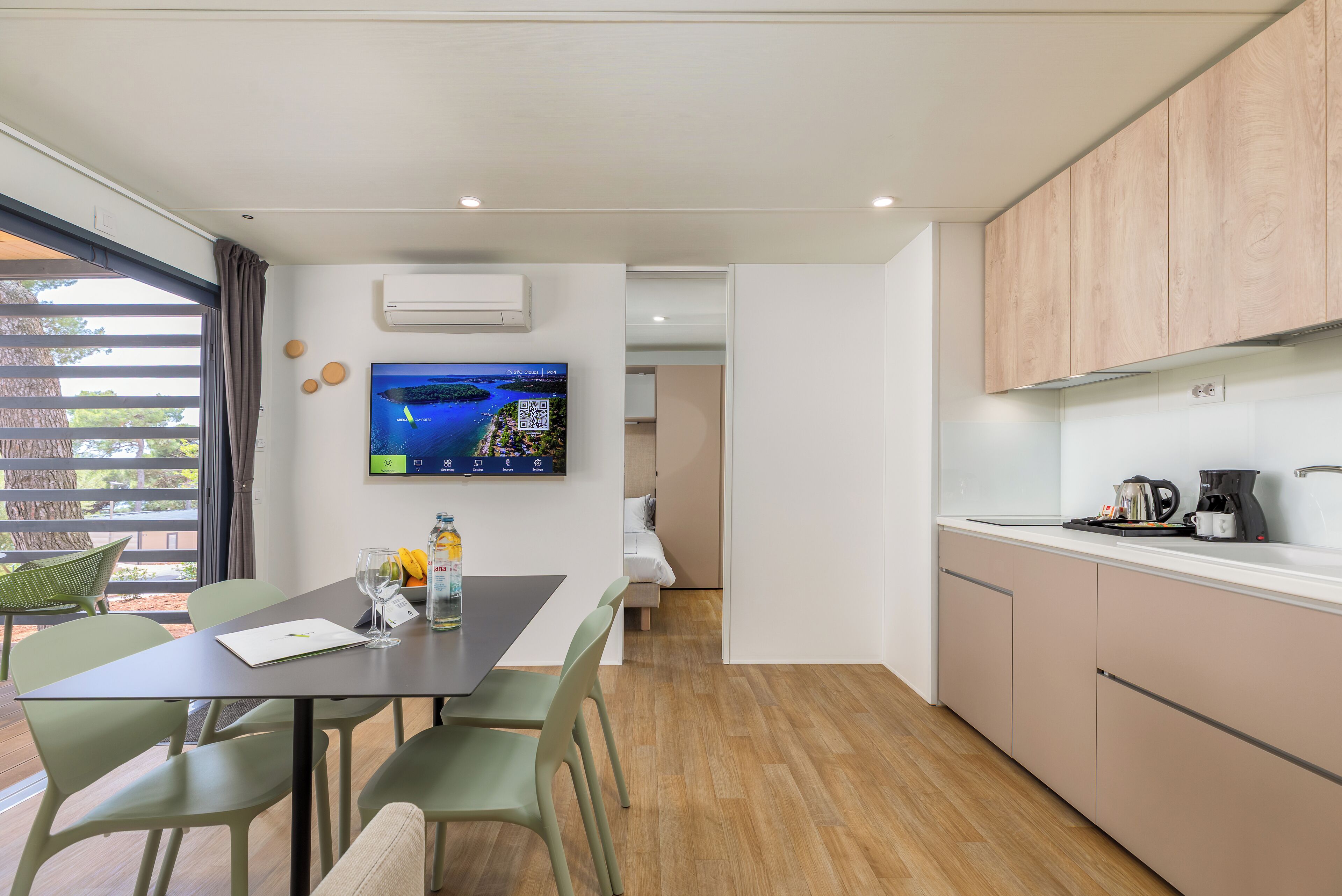 A modern kitchen with a dining area, wooden cabinets, and a flat-screen TV on the wall.