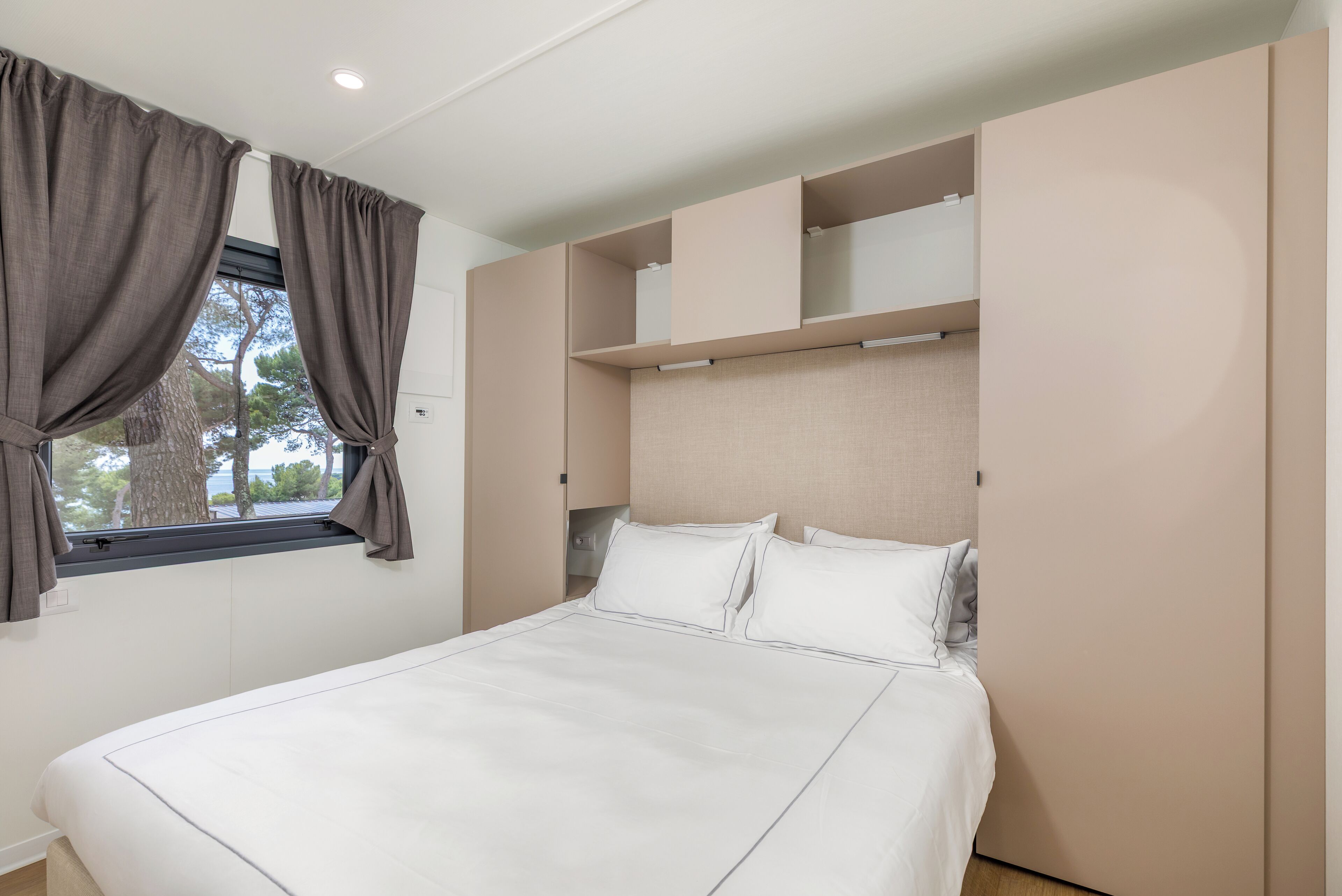 Un lit bien fait, avec du linge de lit blanc, une fenêtre avec des rideaux et des étagères intégrées au-dessus du lit.
