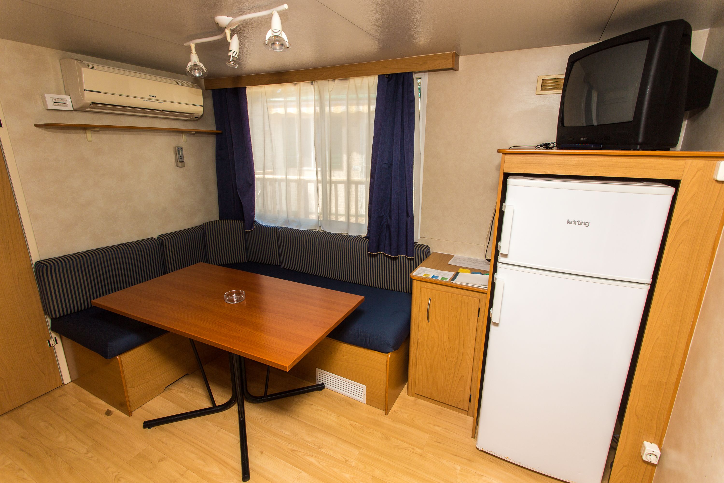 Interior compacto de una caravana con una mesa de madera, asientos tipo banco, una cocina americana con refrigerador y un televisor montado.
