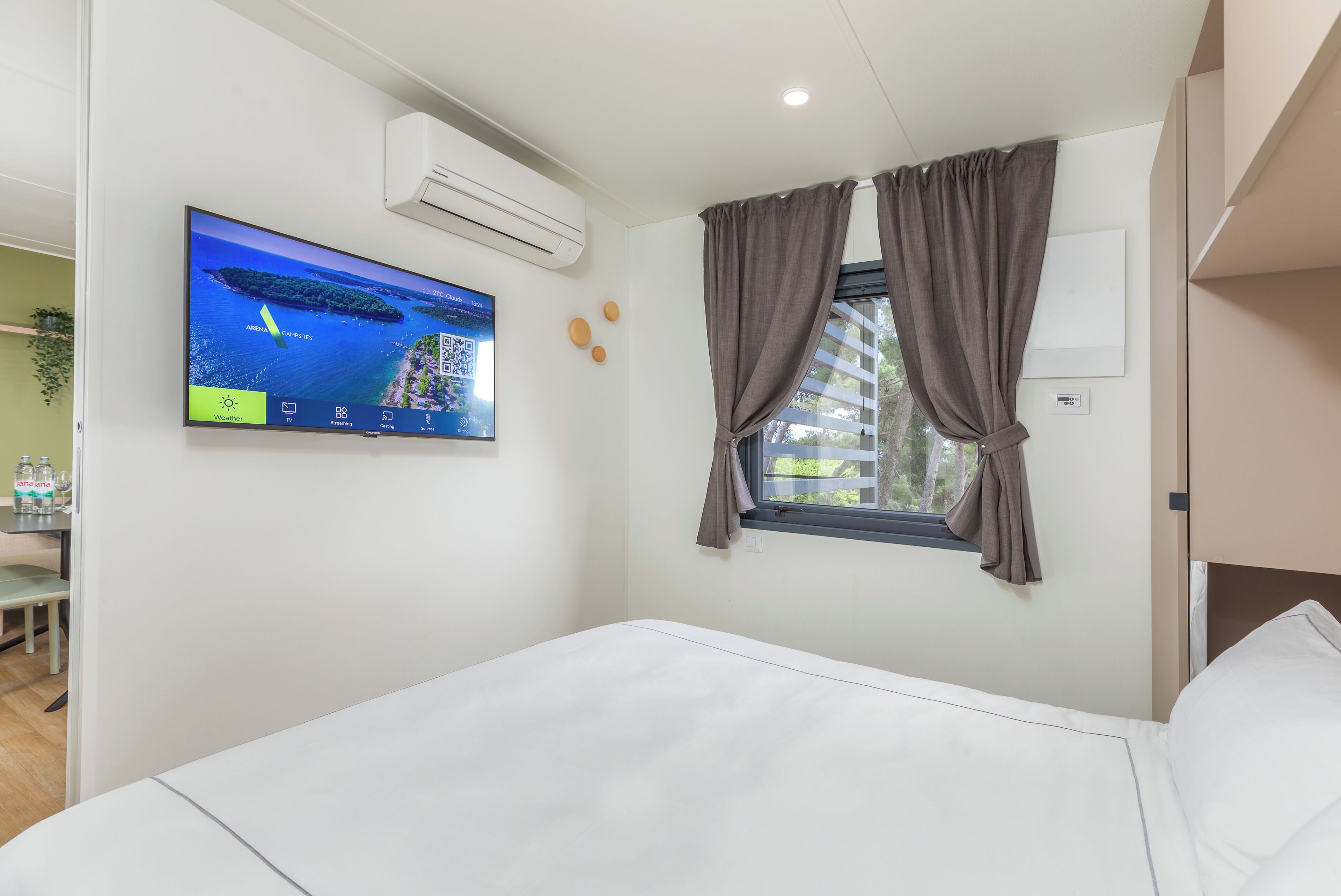 Une chambre d’hôtel avec un lit, un téléviseur fixé au mur, un appareil de climatisation, une fenêtre avec des rideaux et une petite table sur laquelle se trouvent une plante et des bouteilles.