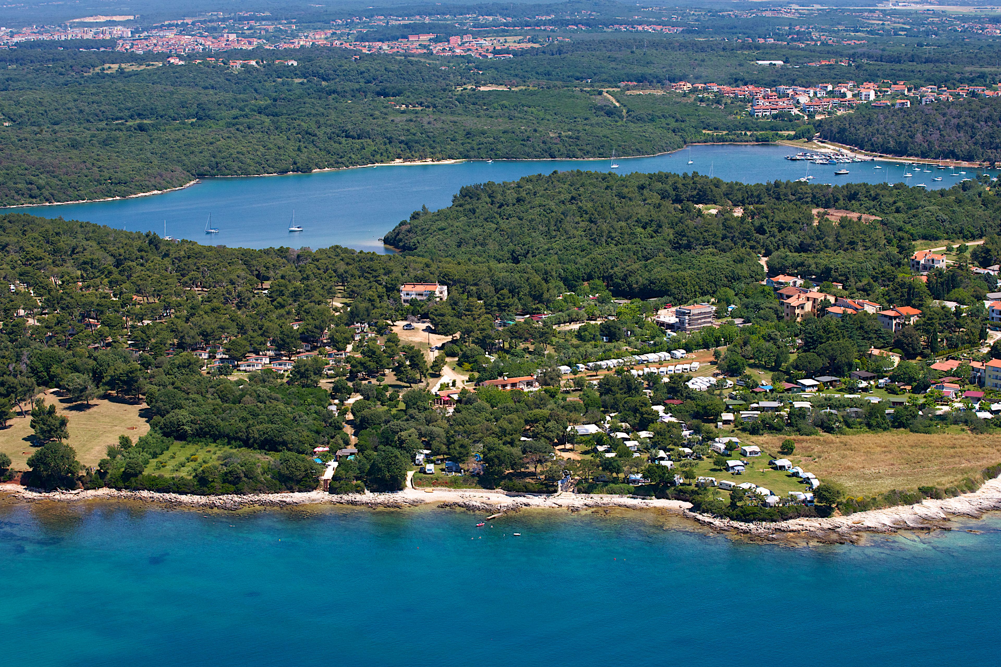 An aerial view of a coastal town with a marina, dense greenery, and a mix of residential and commercial buildings.
