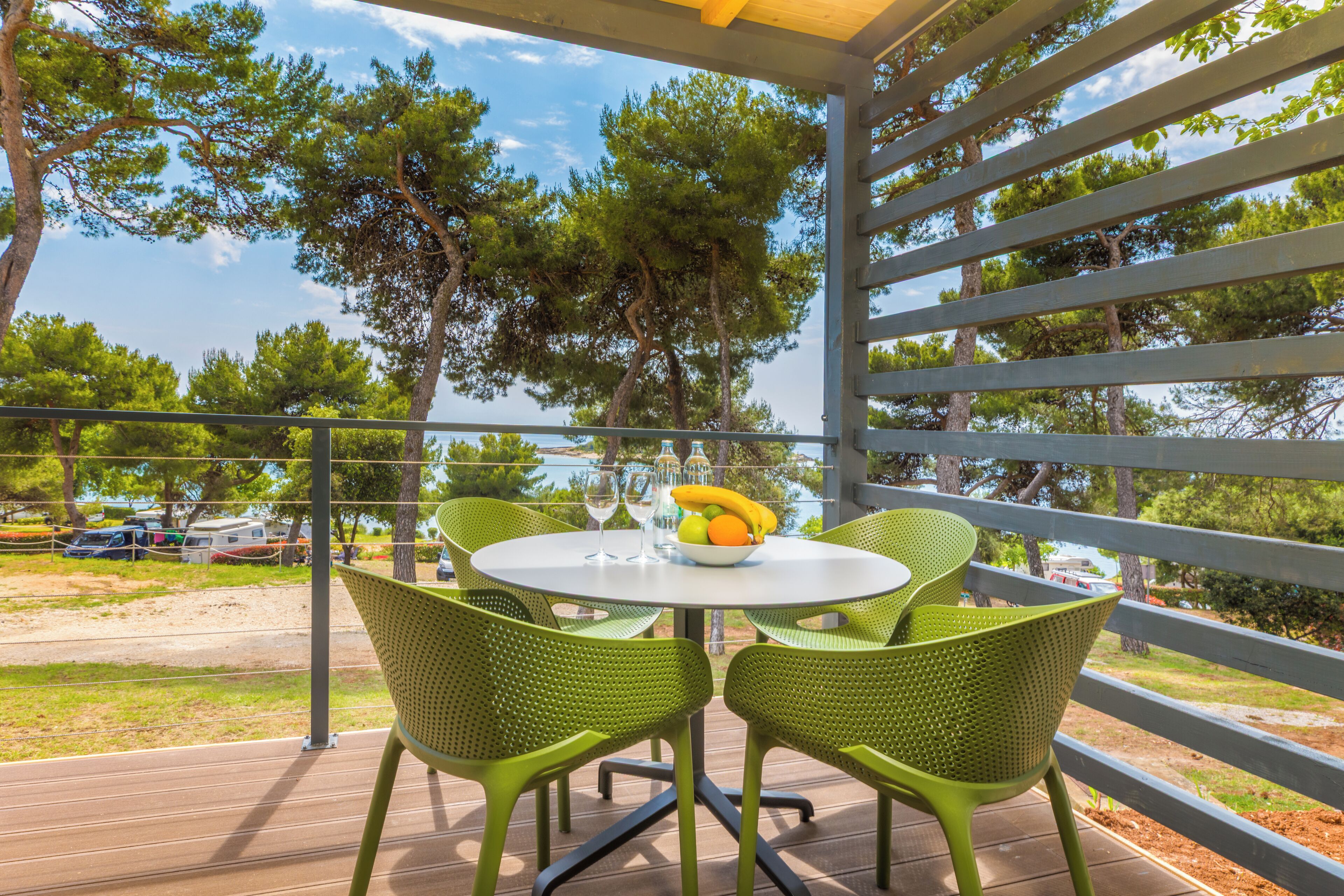 Een overdekt buitengebied met een ronde tafel en groene stoelen, met uitzicht op een strand met geparkeerde voertuigen en bomen.