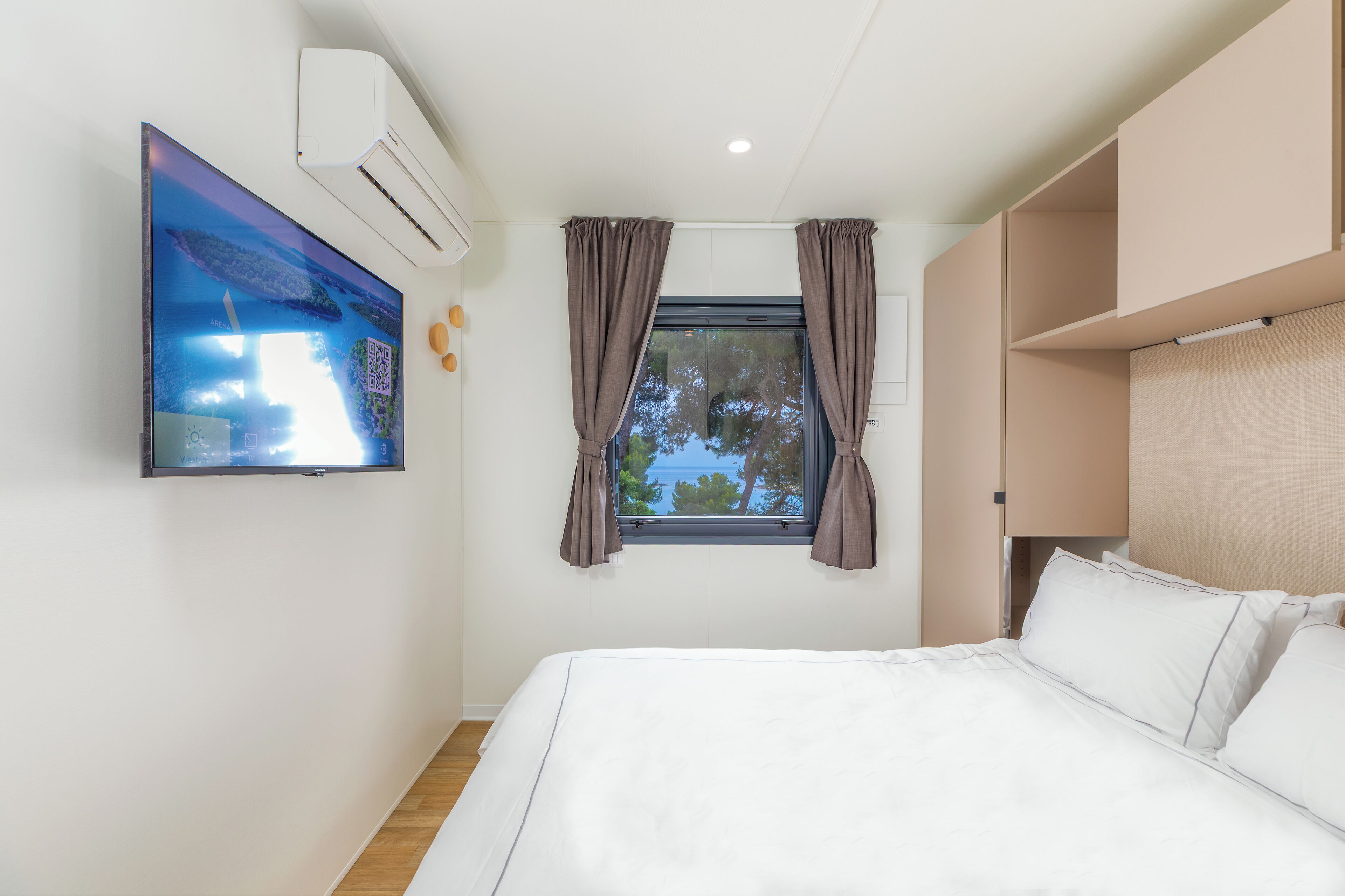Une chambre à coucher comprenant un lit, un téléviseur fixé au mur, un appareil de climatisation et une fenêtre avec des rideaux.