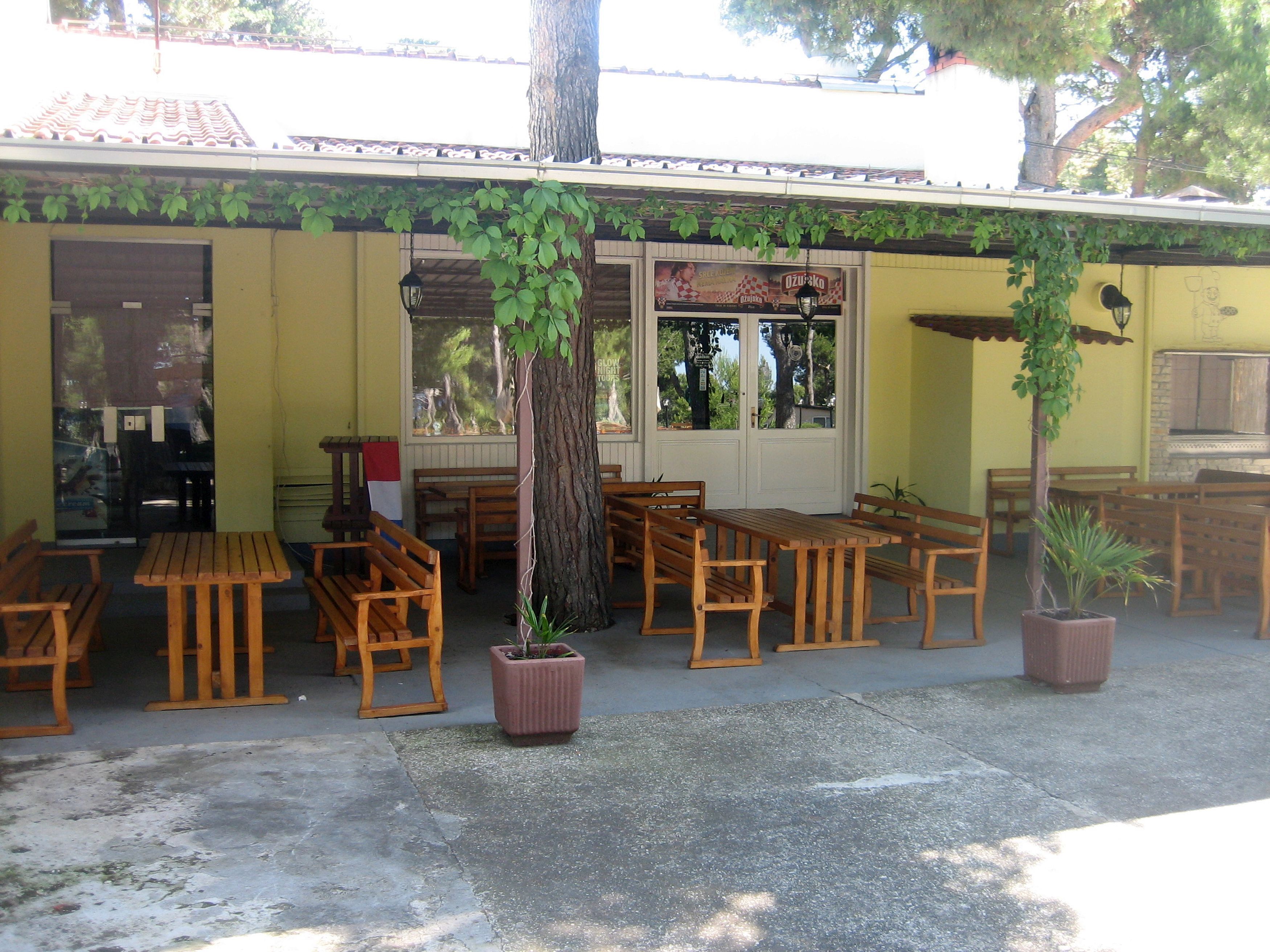 An outdoor cafe with wooden tables and benches, a potted plant, and a tiled roof.
