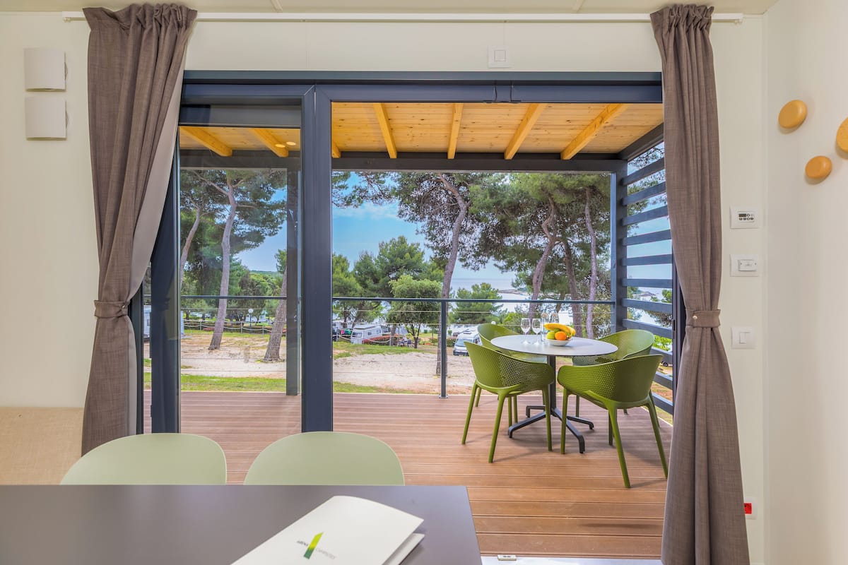 A room with a view of a beach and trees, a table with chairs, and a wooden ceiling.
