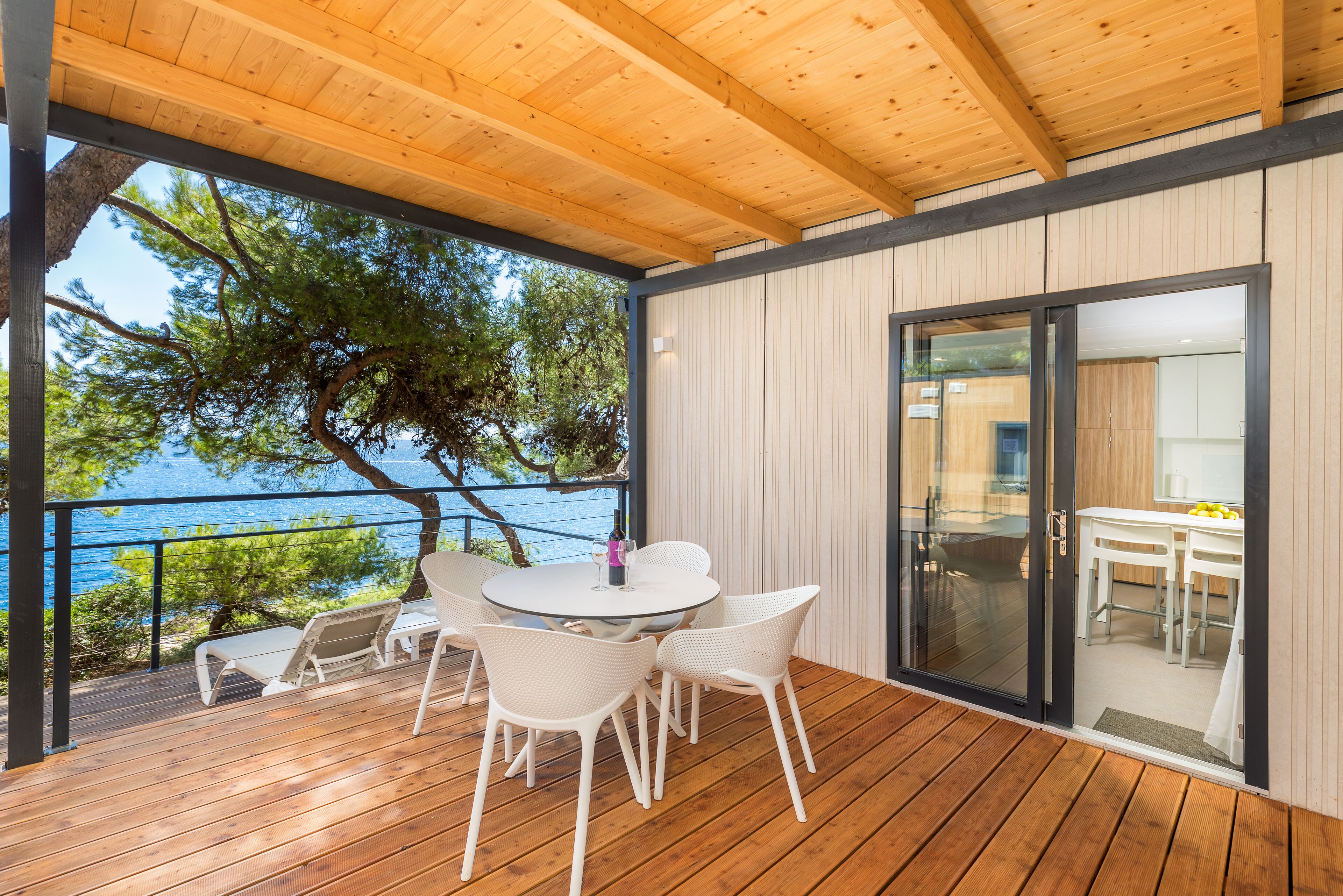 A wooden deck with a round table and chairs, overlooking a body of water and trees.