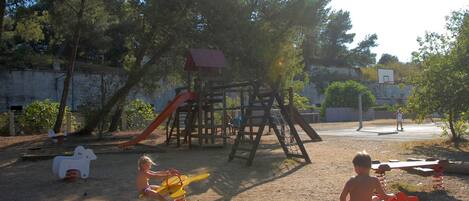 Une aire de jeux dotée de diverses structures, notamment un toboggan et des balançoires, entourée d’arbres et d’un mur en béton.