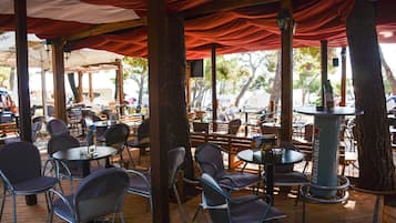 An outdoor seating area with round tables and chairs, covered by red fabric canopies, and surrounded by trees.