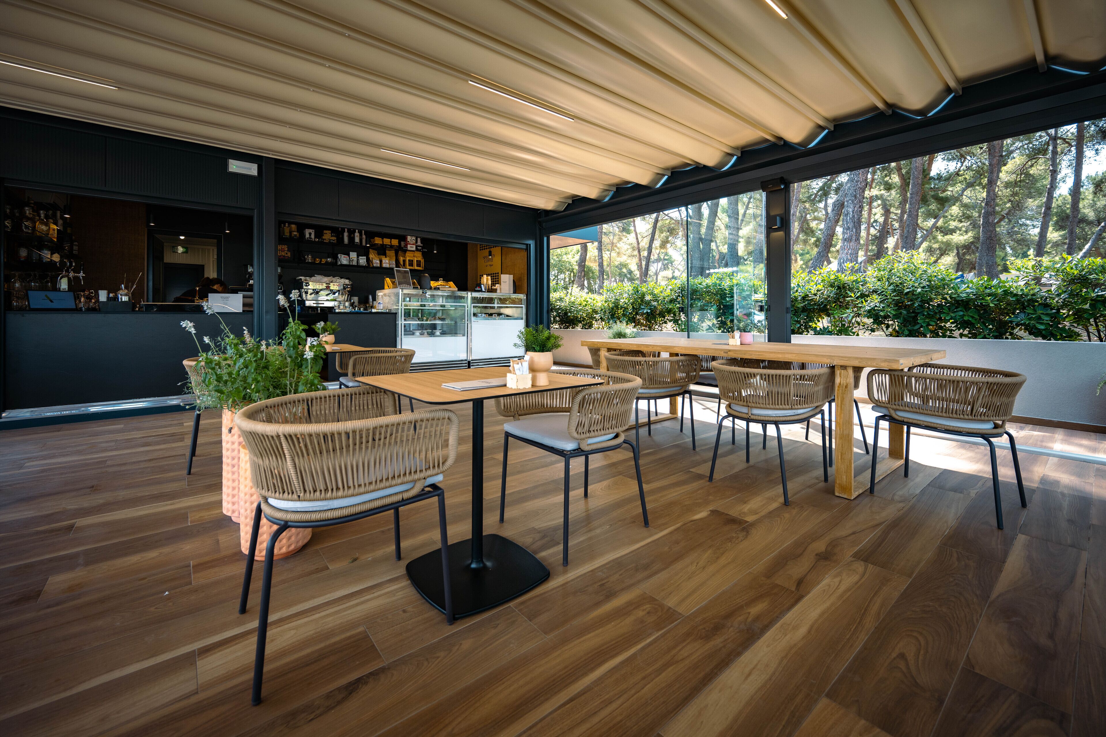 A modern outdoor seating area with wooden tables and wicker chairs, surrounded by large windows offering a view of trees.