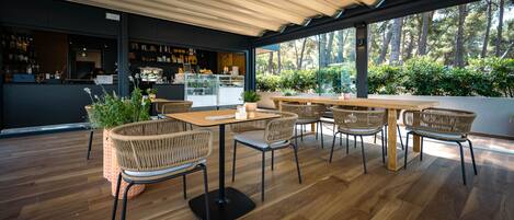 A modern outdoor seating area with wooden tables and wicker chairs, surrounded by large windows offering a view of trees.