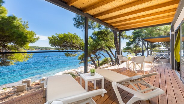 A wooden deck with lounge chairs and a small table, overlooking a clear blue sea and a tree-lined shoreline. - Arena Stoja Camping Homes (Pula)