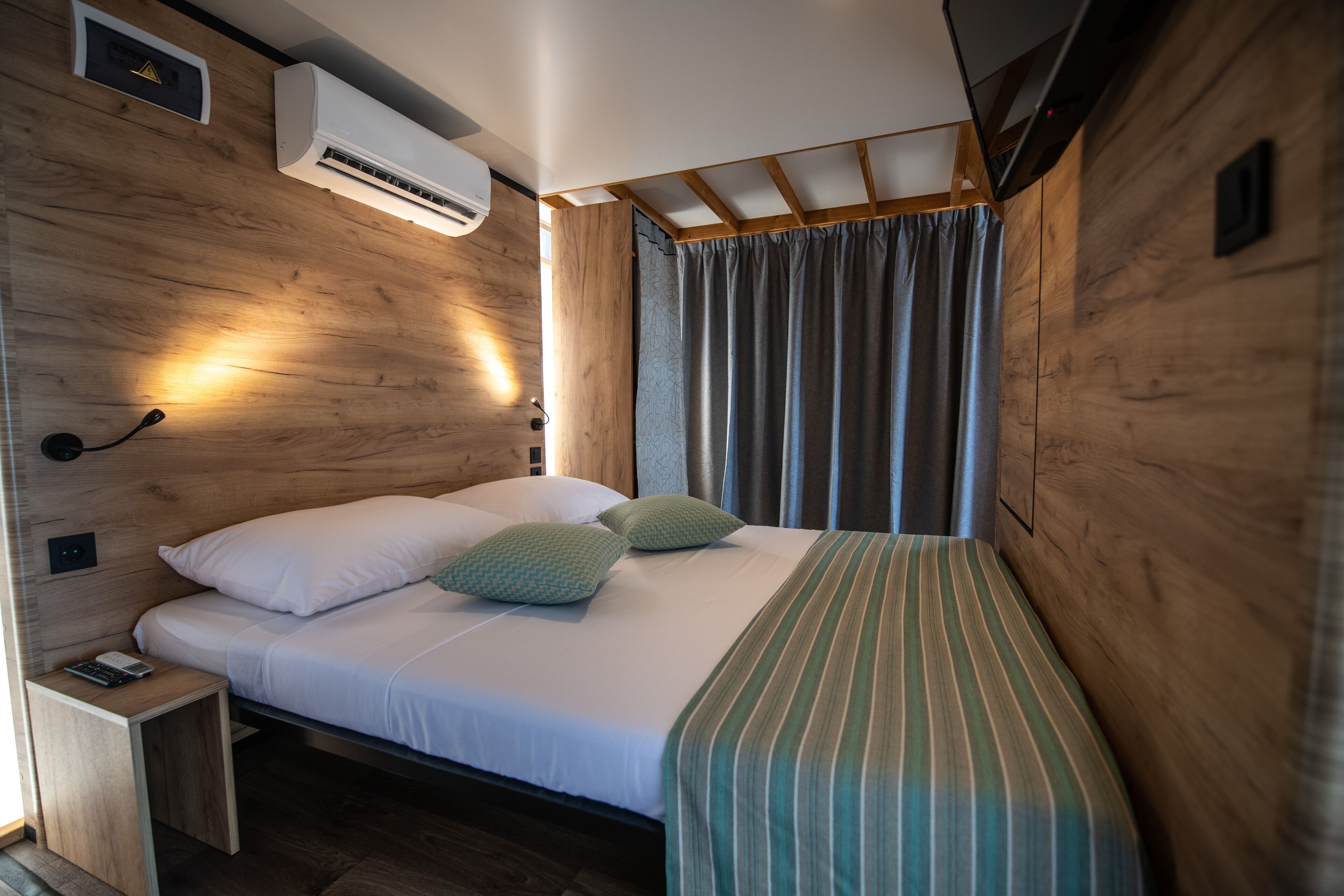 A compact bedroom with a wooden wall, a bed with white bedding, a striped blanket, and a small bedside table.