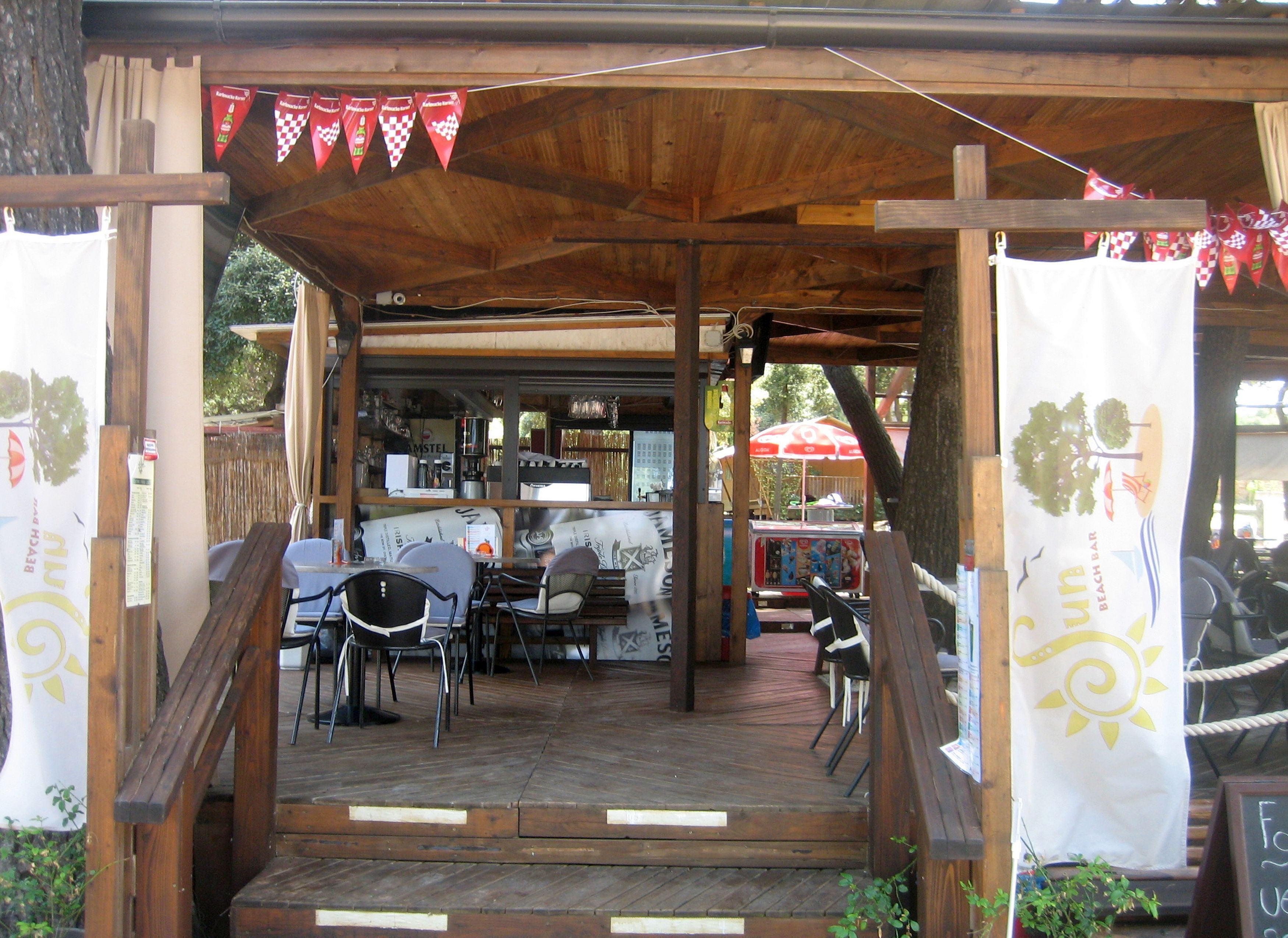 Een houten constructie buiten met tafels en stoelen, een overdekte zithoek en zicht op een bar op de achtergrond.