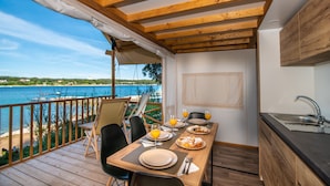 A dining area with a view of the sea, wooden beams, a table set for two, and a small kitchen area. - Arena Stoja Camping Homes (Pula)