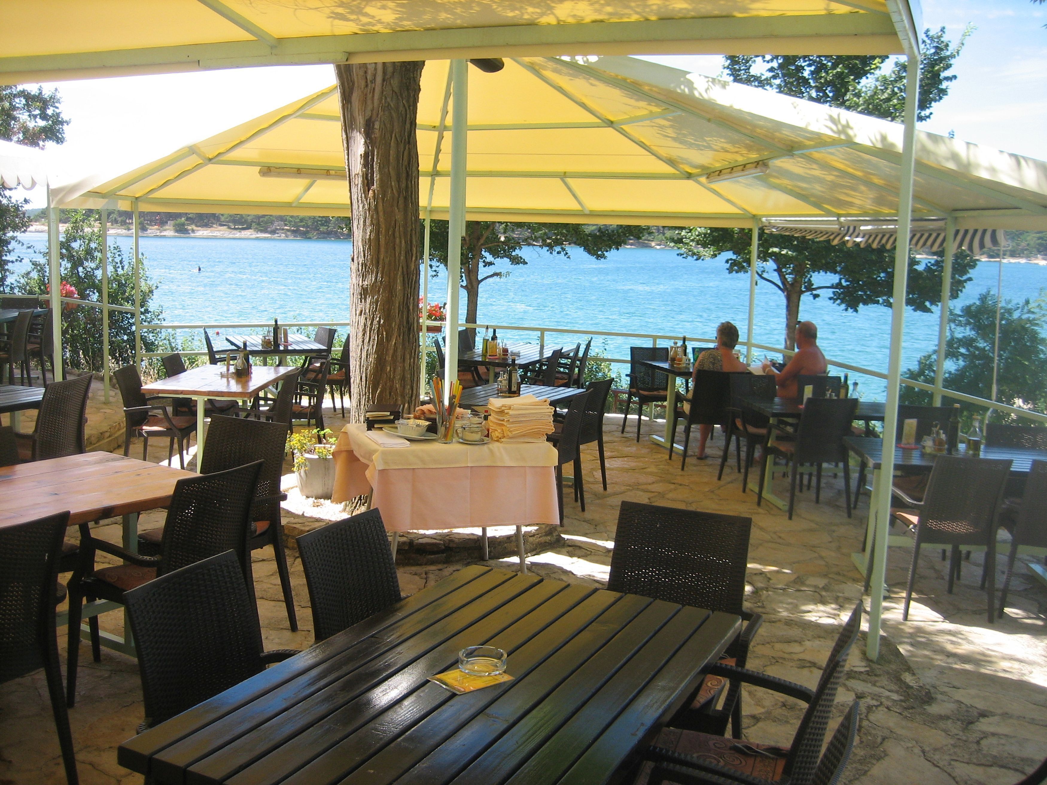 An outdoor dining area with tables and chairs, a large umbrella, and a view of the sea.