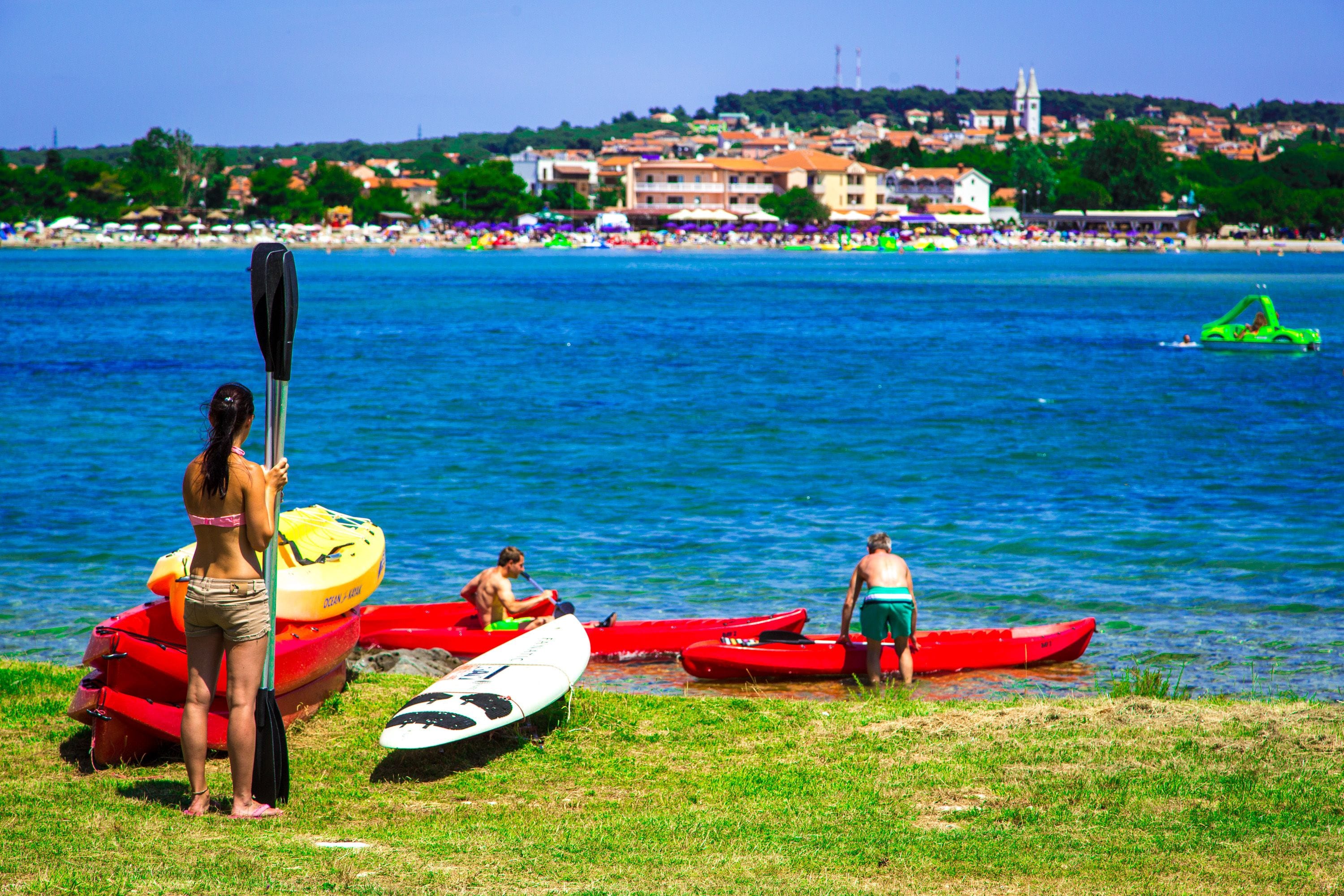 A beach with kayaks and a paddleboard, a person standing with a paddle, and a town in the background.