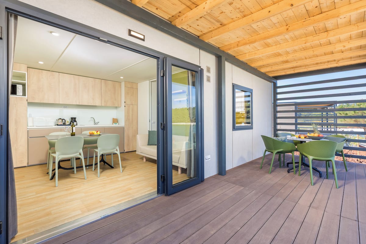 A modern kitchen with wooden cabinets, a dining area with green chairs, and a wooden deck with a view of the outdoors.