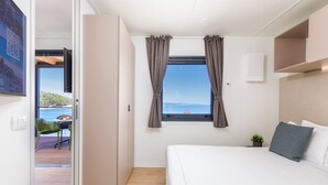 A bedroom with a bed, a window with curtains, a small plant, and a view of the sea. - Arena Stupice Camping Homes (Medulin)