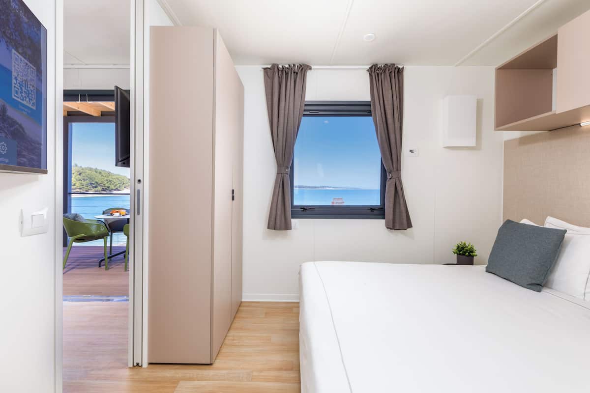 A bedroom with a bed, a window with curtains, a small plant, and a view of the sea.