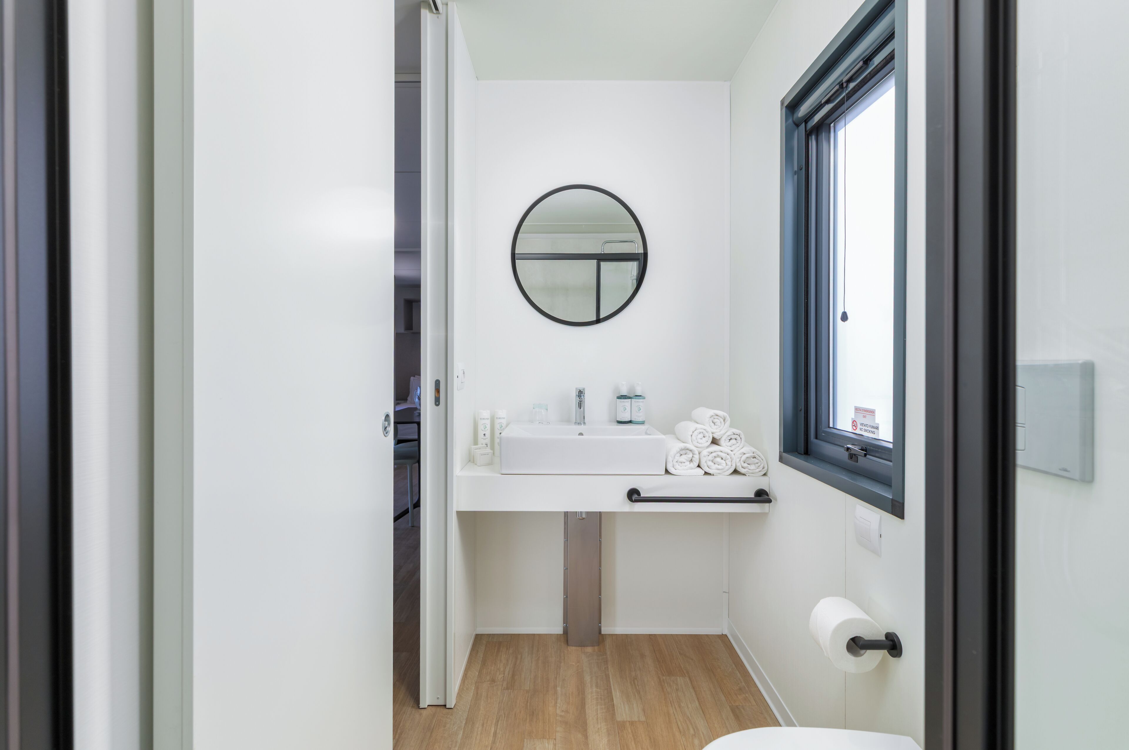 A modern bathroom with a white sink, a round mirror, and a window.